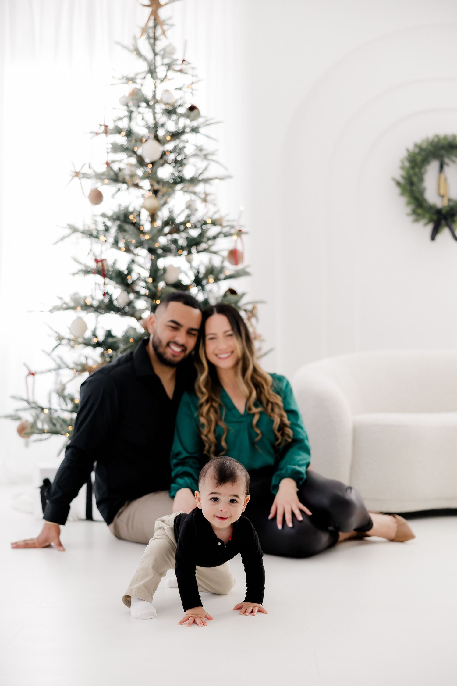 Family posing for holiday portrait in front of decorated Christmas tree in white room.