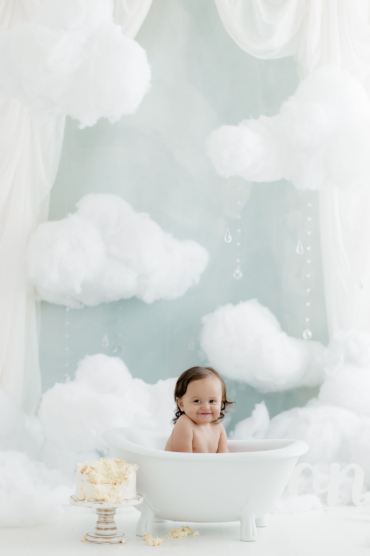 Happy baby smiles in white clawfoot tub with smash cake beside fluffy clouds in blue studio.