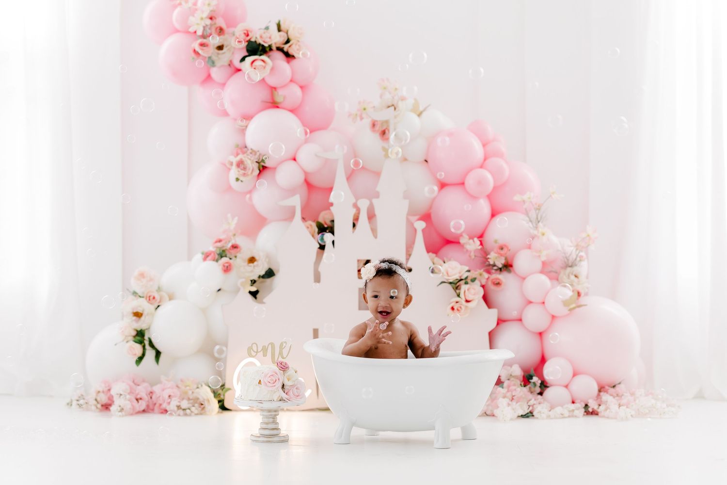 Pink and white balloon arch with floral decorations against a white backdrop creates an elegant party setting.