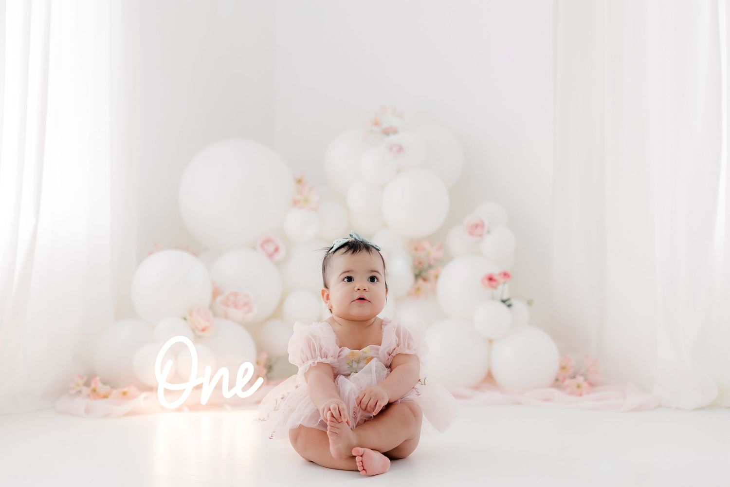 Baby in pink outfit sits in front of illuminated 'One' sign and white balloon backdrop.