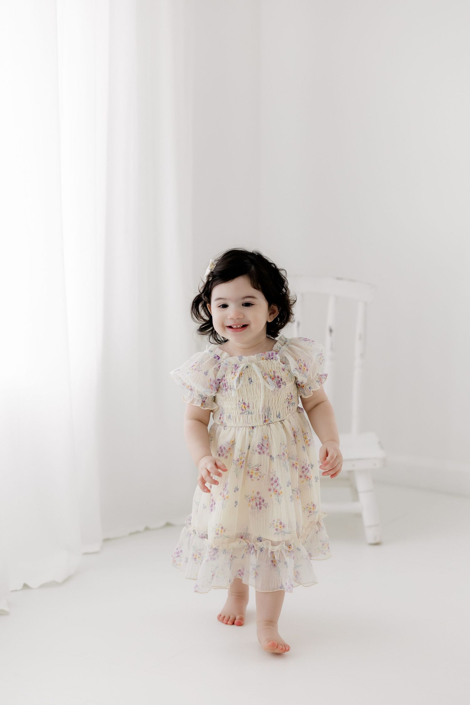 Toddler girl in floral dress smiling and walking barefoot in bright white studio setting.