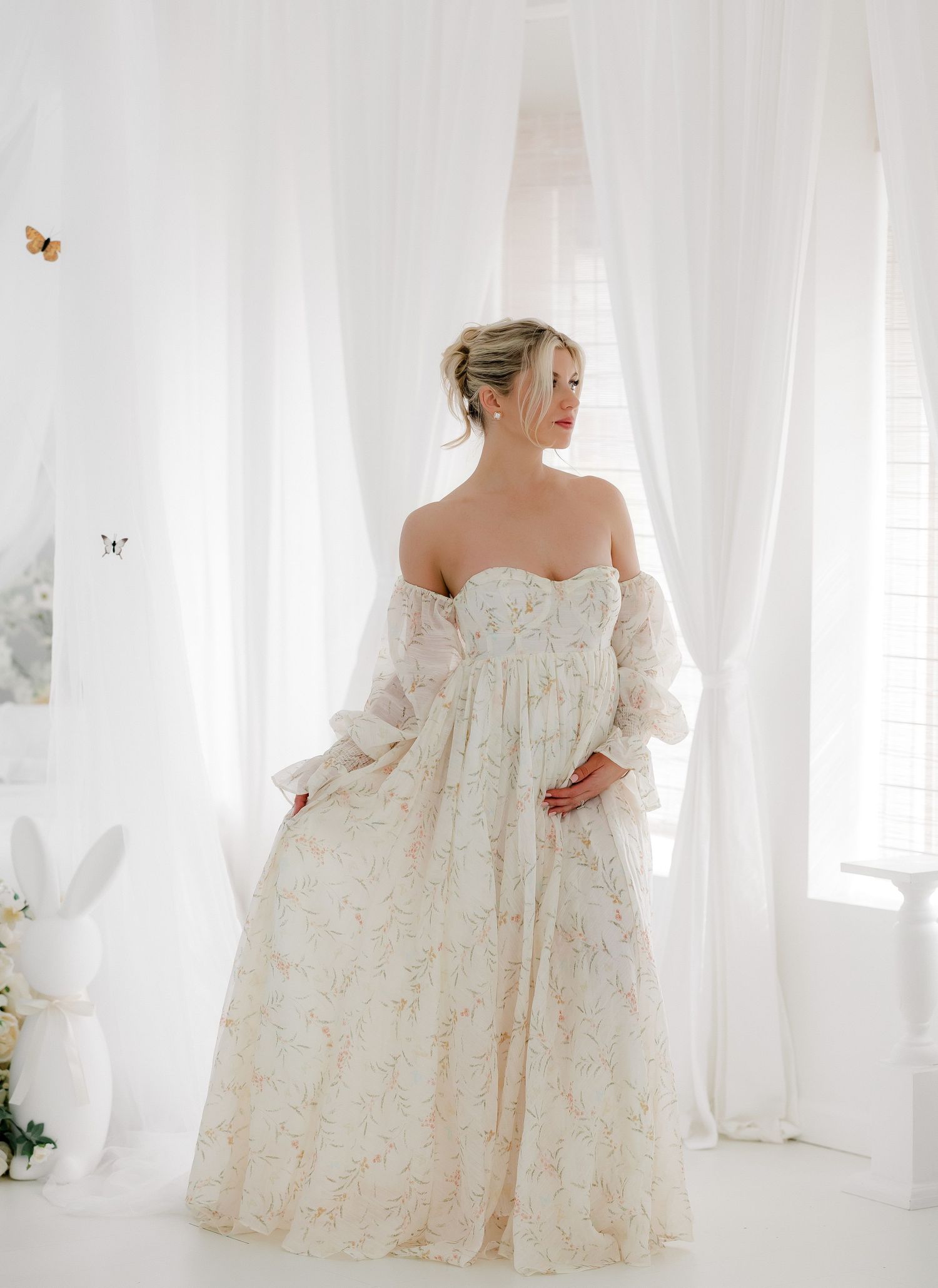 A wedding dress with delicate lace details and off-shoulder design is displayed in a bright white room with sheer curtains.