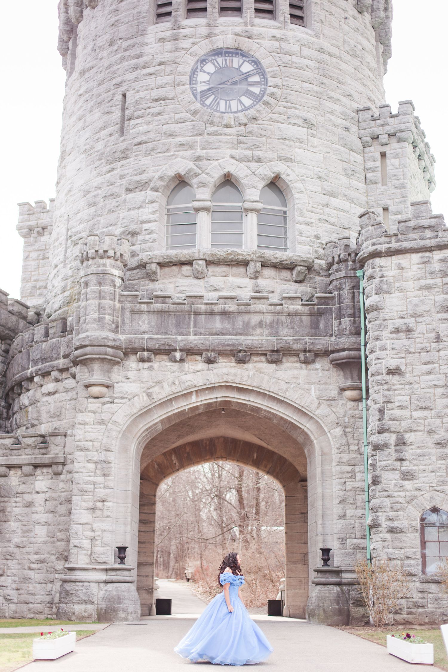 A panoramic view of a stone castle entrance with arched doorways and clock tower shows a blue gown against the gray walls.