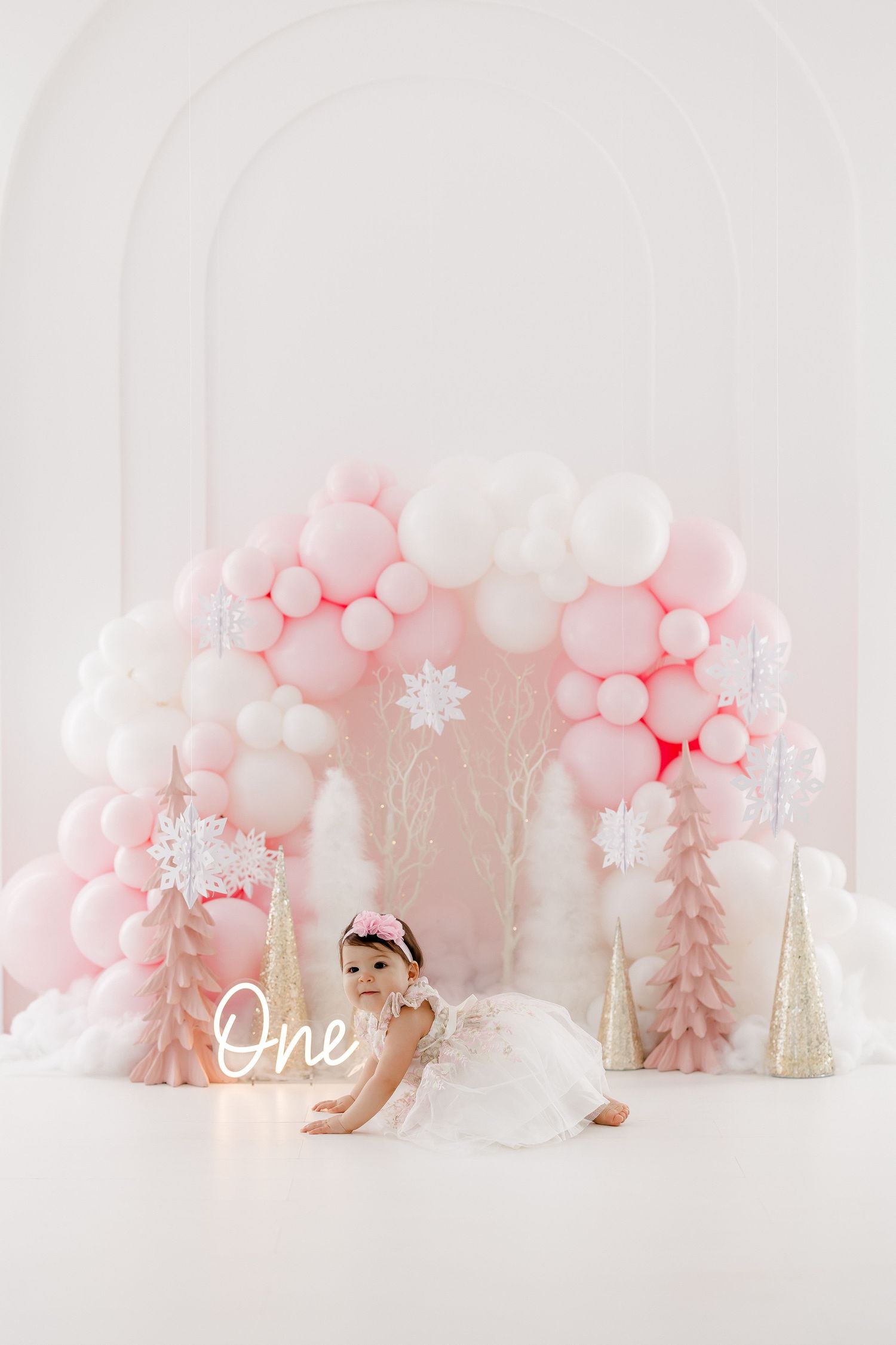 Pink and white winter wonderland cake smash setup with balloons, snowflakes, and trees for first birthday celebration.