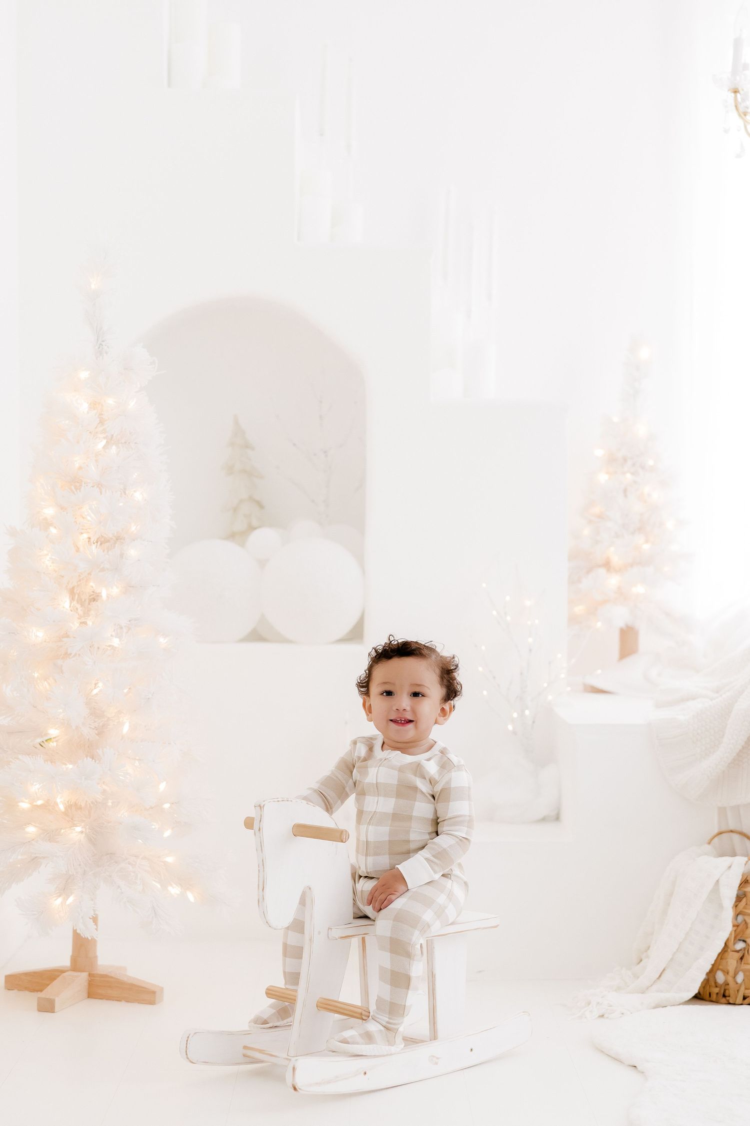 Baby in white outfit plays with Christmas decorations in bright minimalist holiday setting.