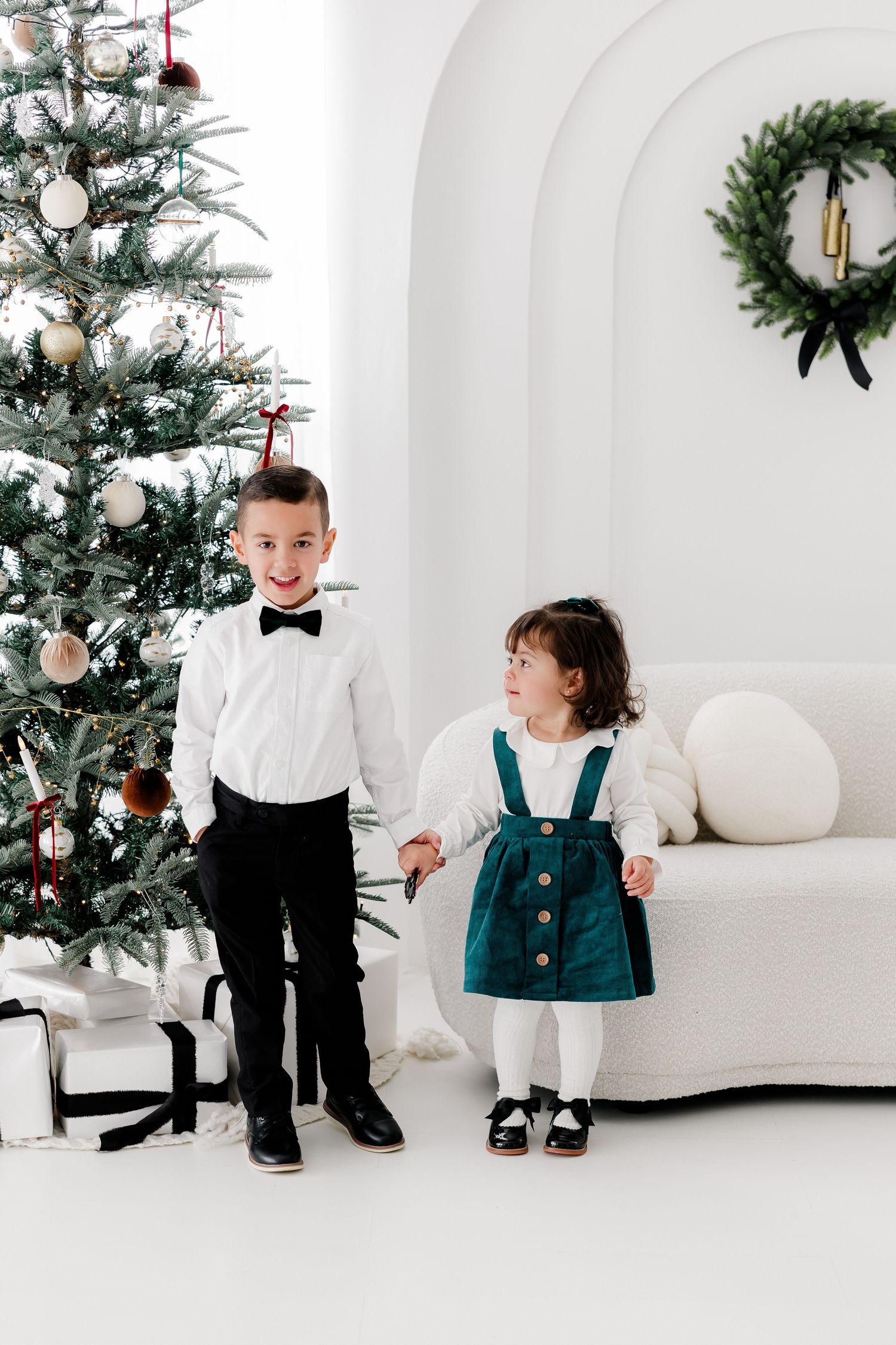Two children in formal attire stand beside decorated Christmas tree in white room.nj holiday mini sessions