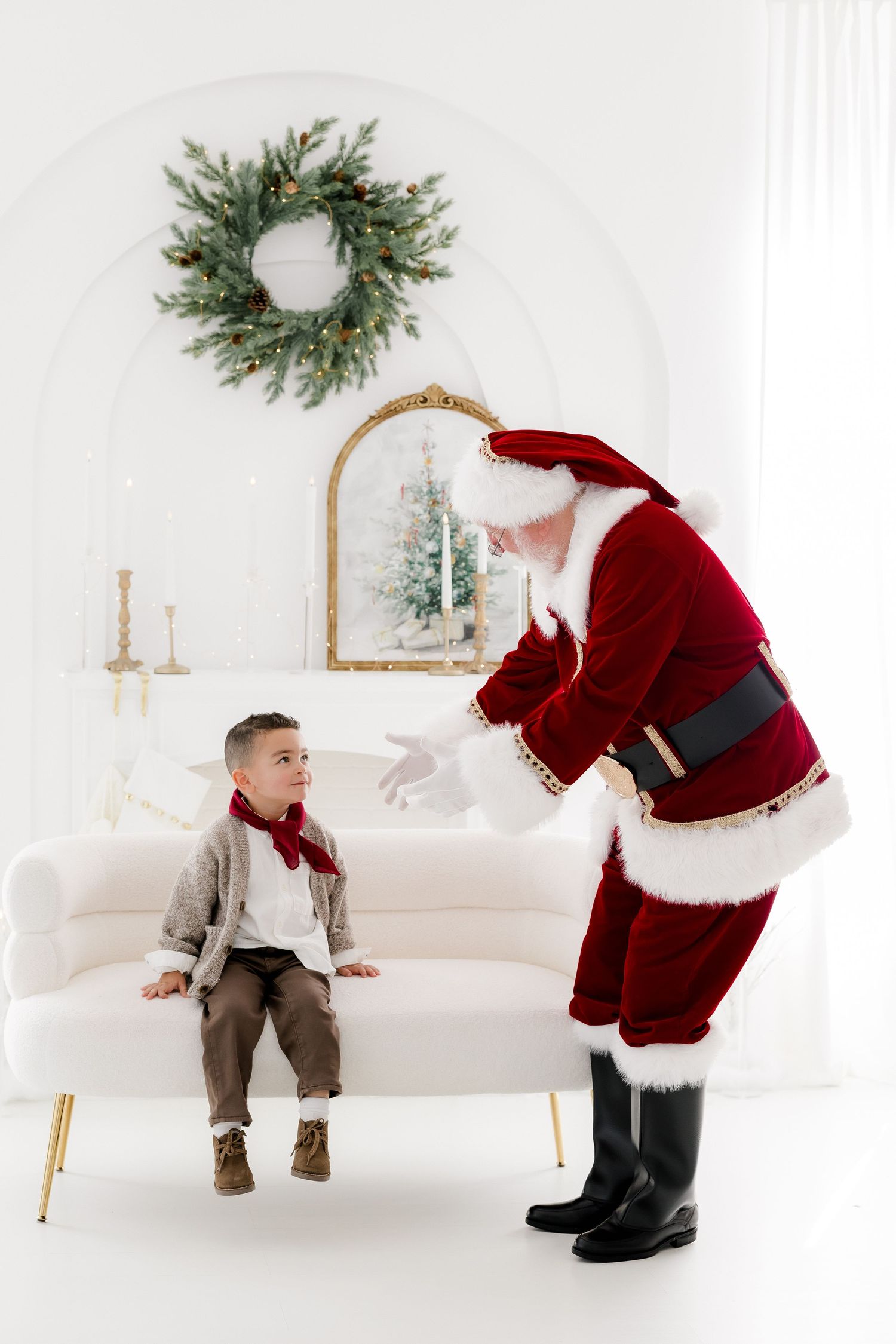 Santa Claus leaning toward a young boy seated on a pink velvet sofa in a white Christmas decorated room.