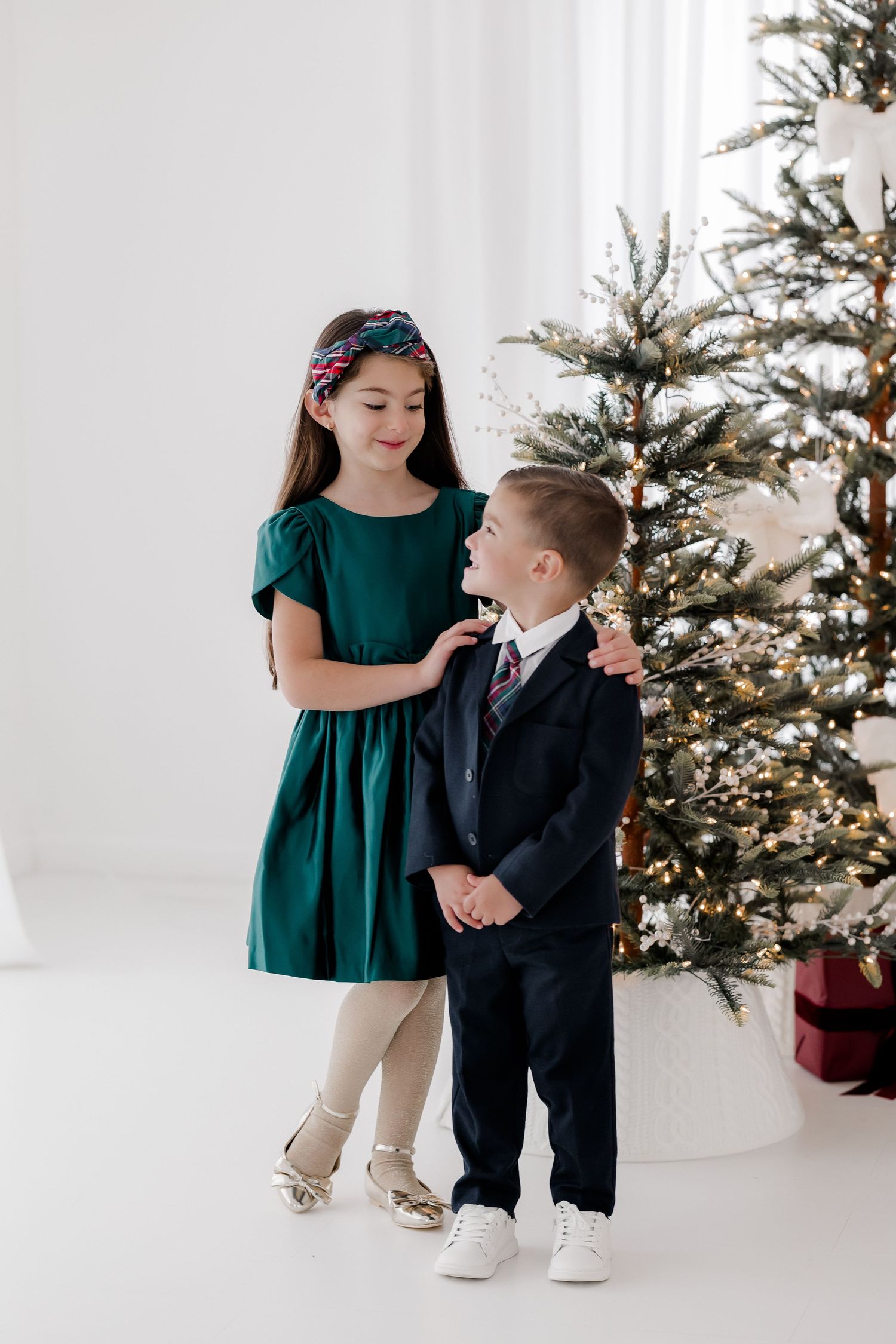 Two children in holiday attire pose by a decorated Christmas tree, girl in green dress and boy in navy suit.