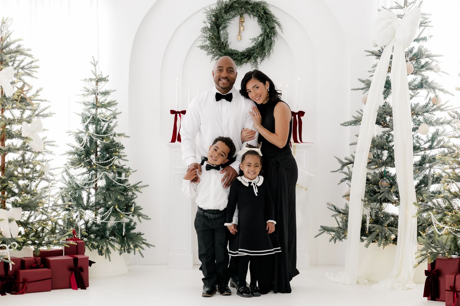 Family of four in elegant black and white outfits posing by Christmas trees and gifts in a white studio.
