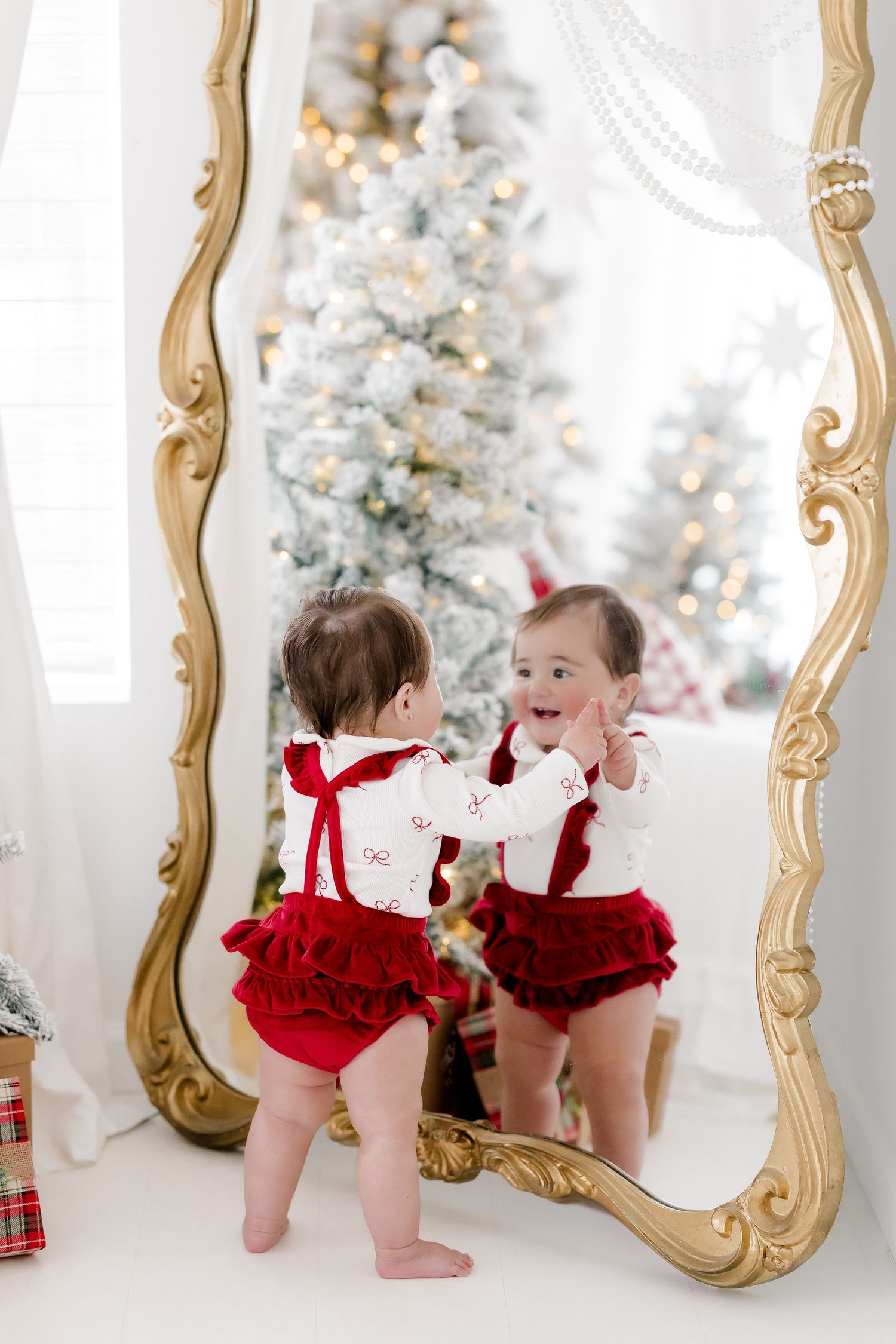 Baby girl in red ruffled bloomers smiling at her reflection in a gold mirror with a decorated Christmas tree behind her.