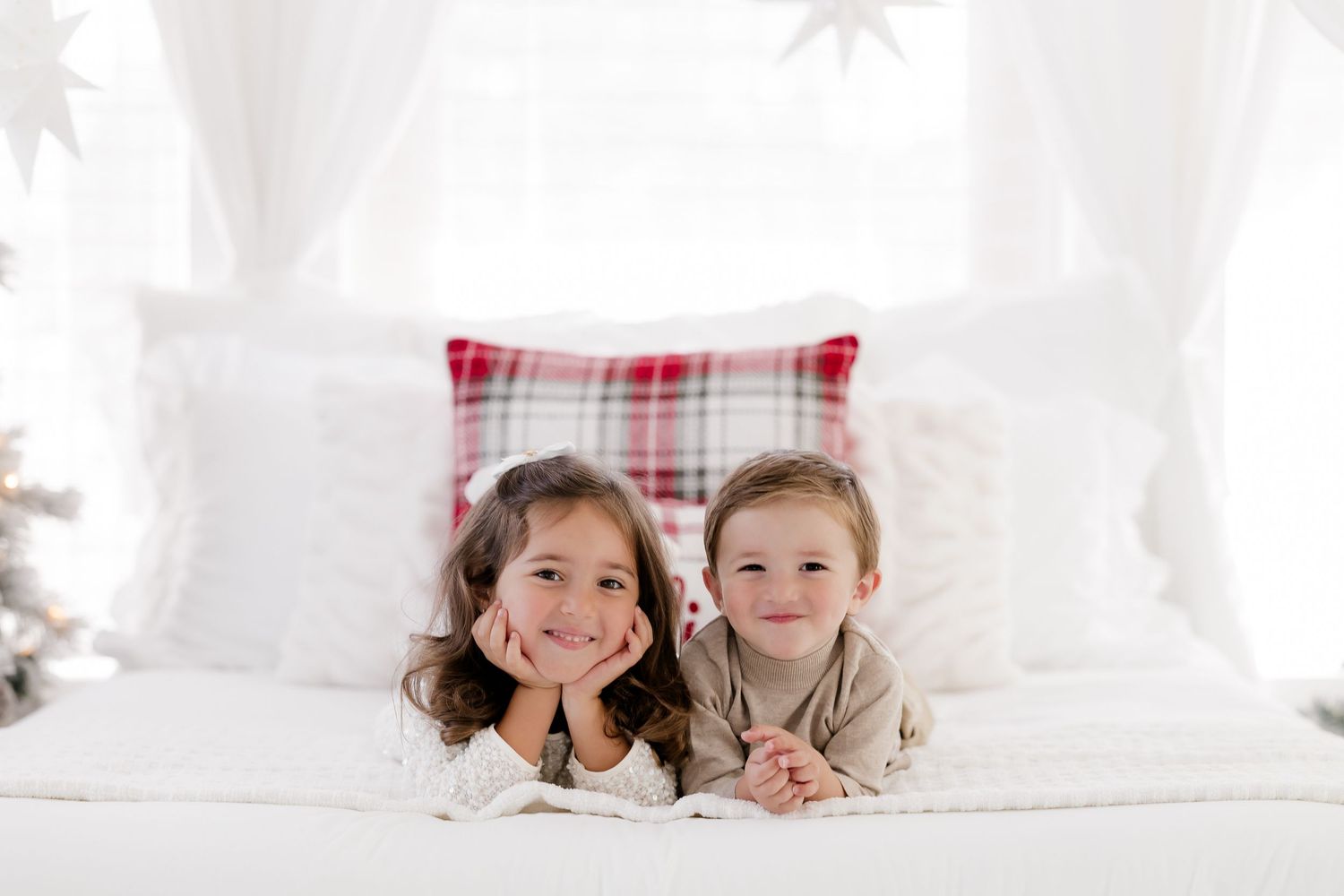 Two young children lying on a white bed with a red plaid pillow, smiling at camera in a festive holiday setting.