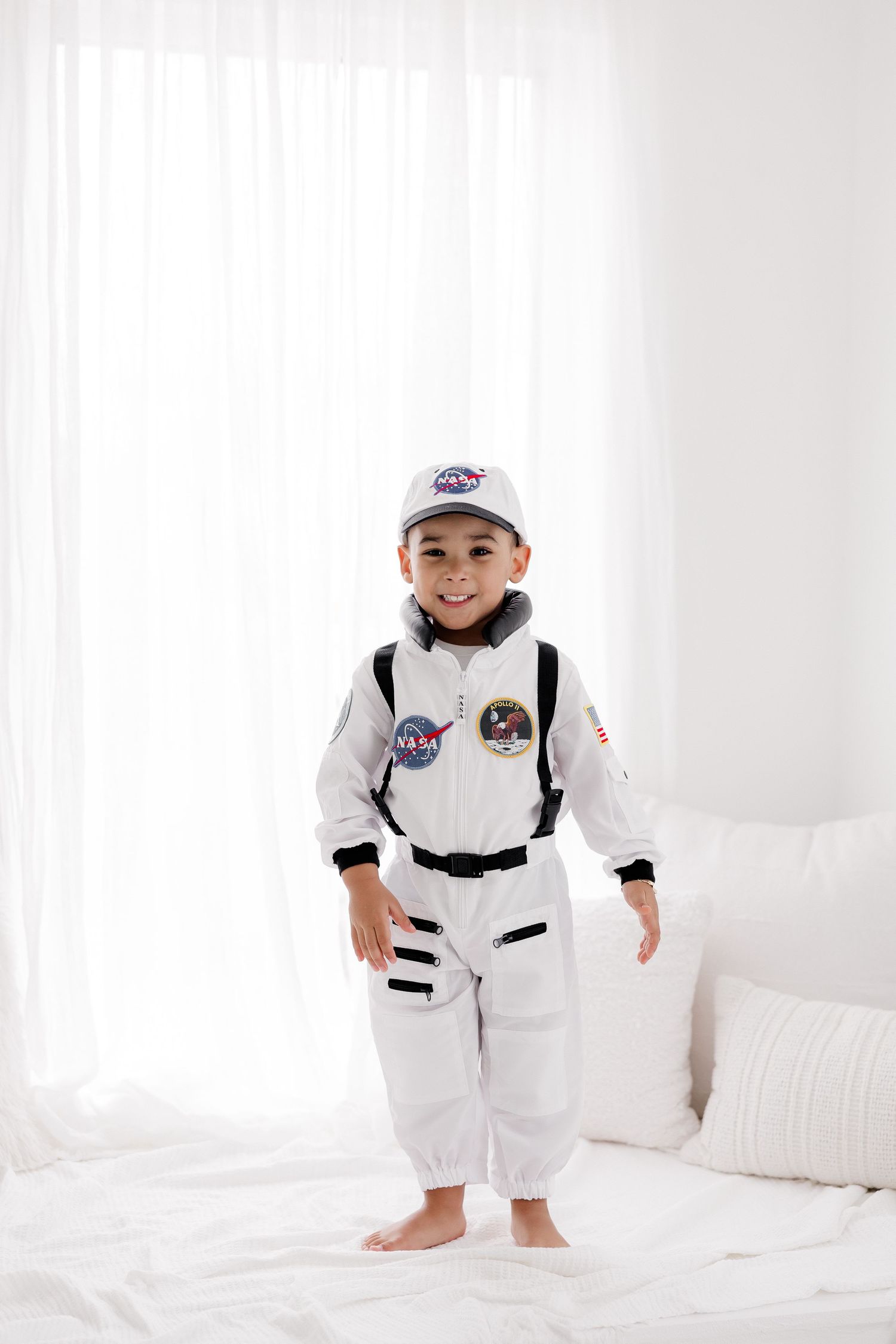 A child wearing a white NASA astronaut costume and baseball cap poses on a white couch in four different positions.