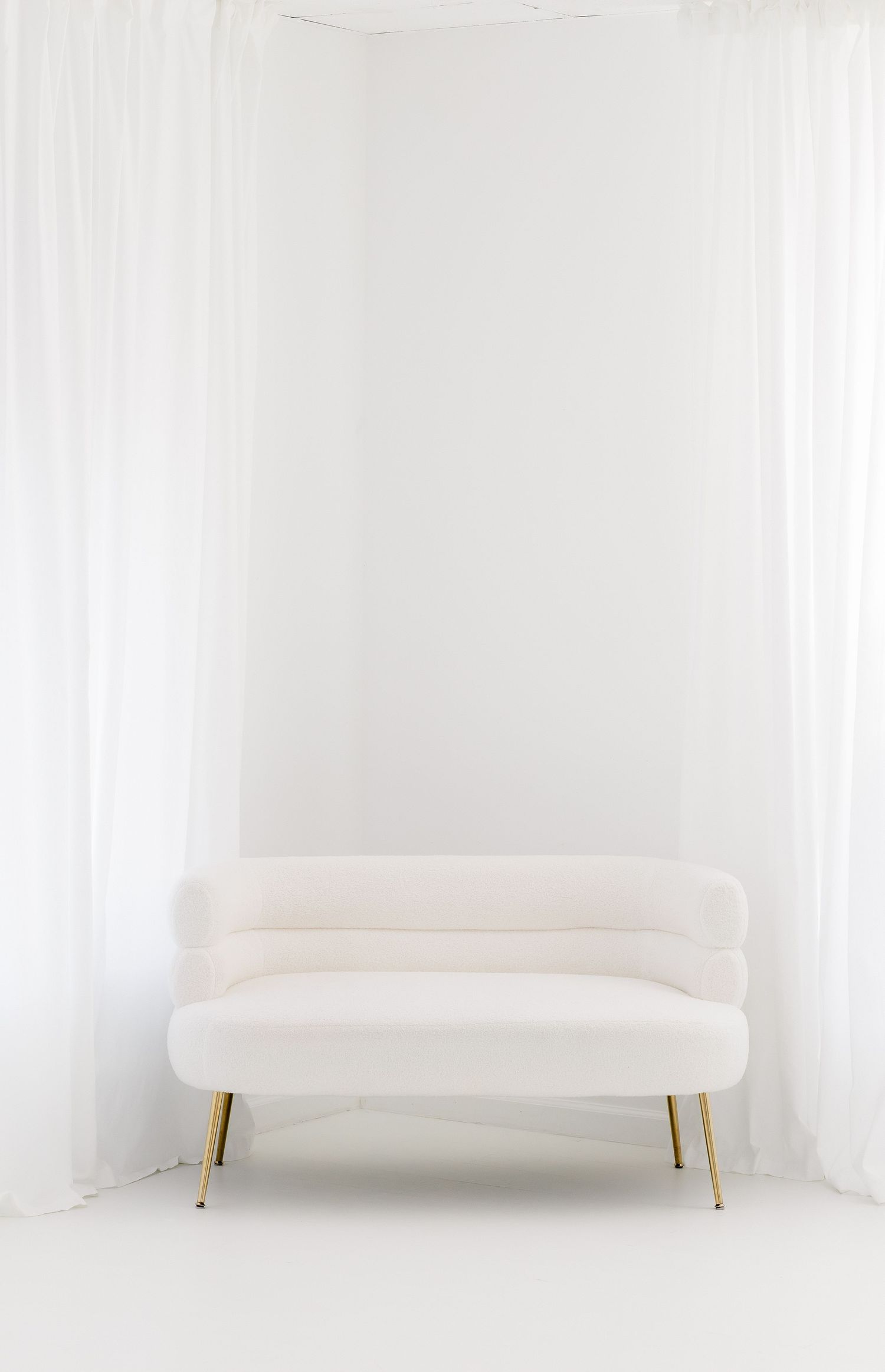 Minimalist cream-colored loveseat with gold legs sits against white curtains in an all-white contemporary setting.