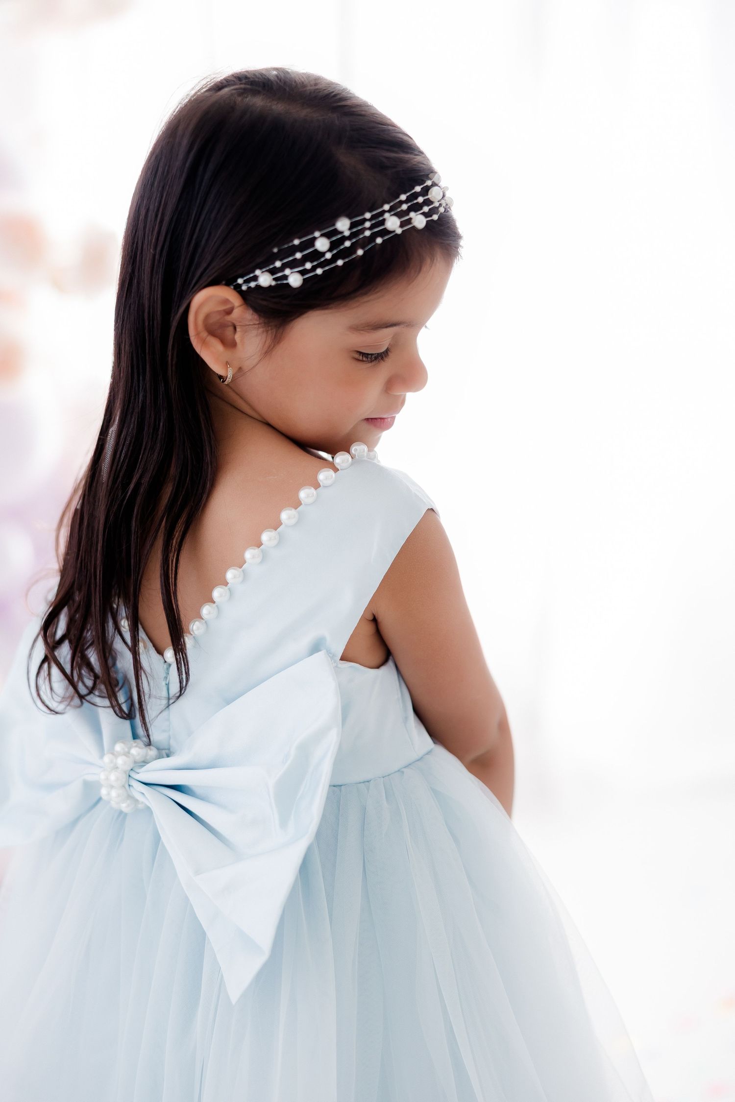 Young girl in light blue tulle dress with pearl bow and headband, looking down gracefully.