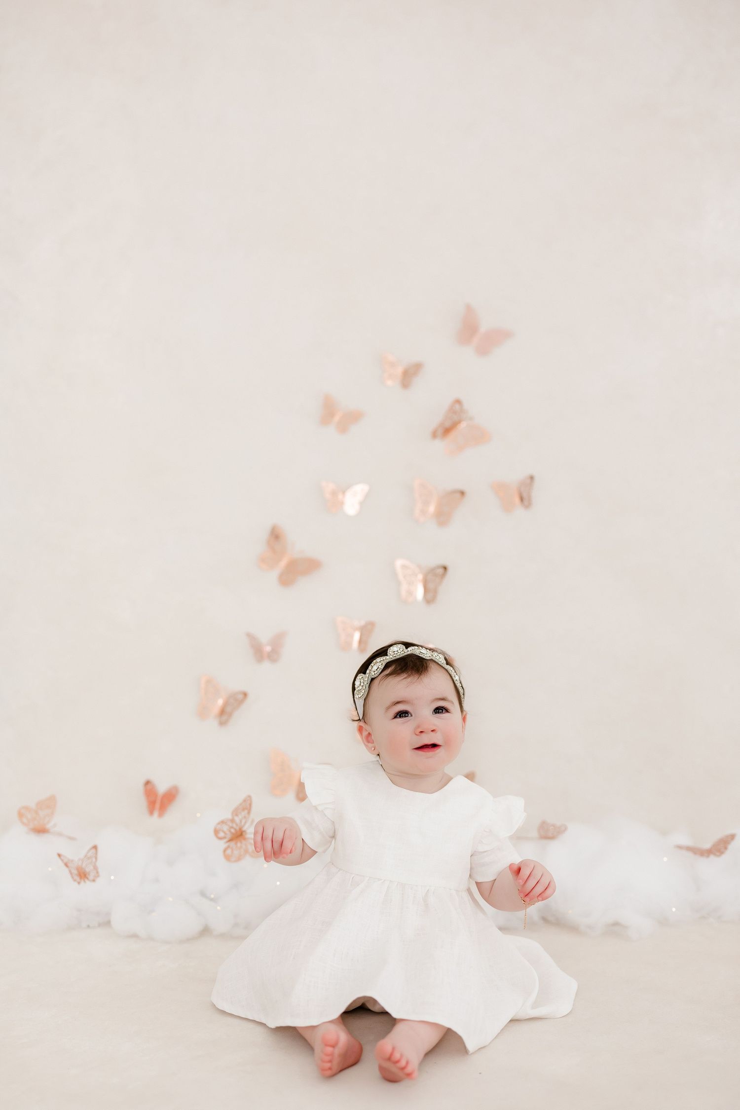 A baby in a white dress sits on a cream backdrop with delicate pink butterflies floating around them.