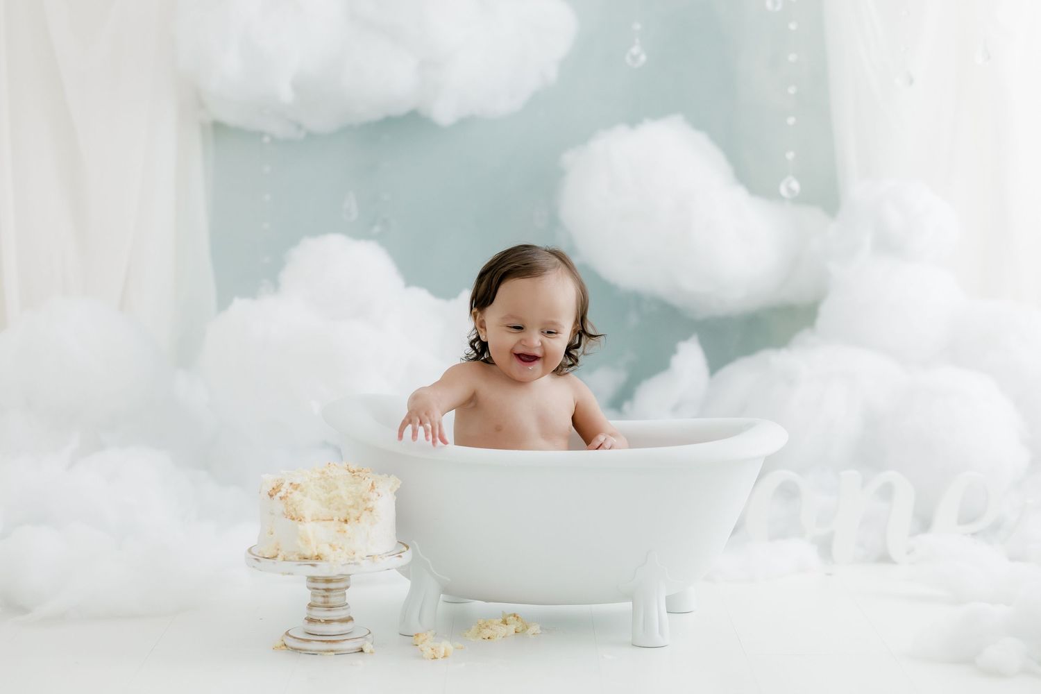 Smiling baby sitting in a white clawfoot bathtub surrounded by fluffy clouds in a dreamy photo studio setup.