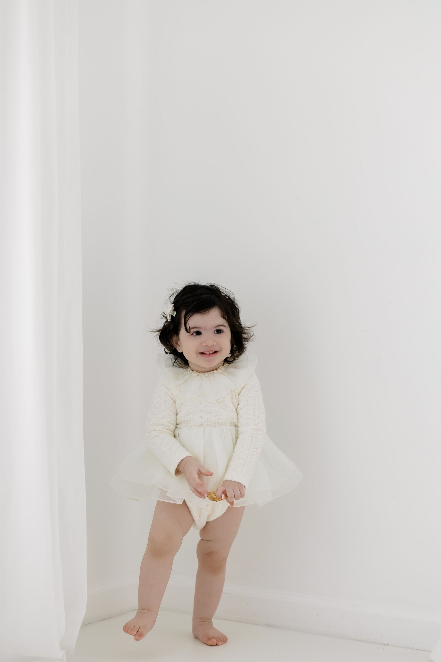 Smiling toddler girl in white long-sleeve dress standing barefoot against a white wall.