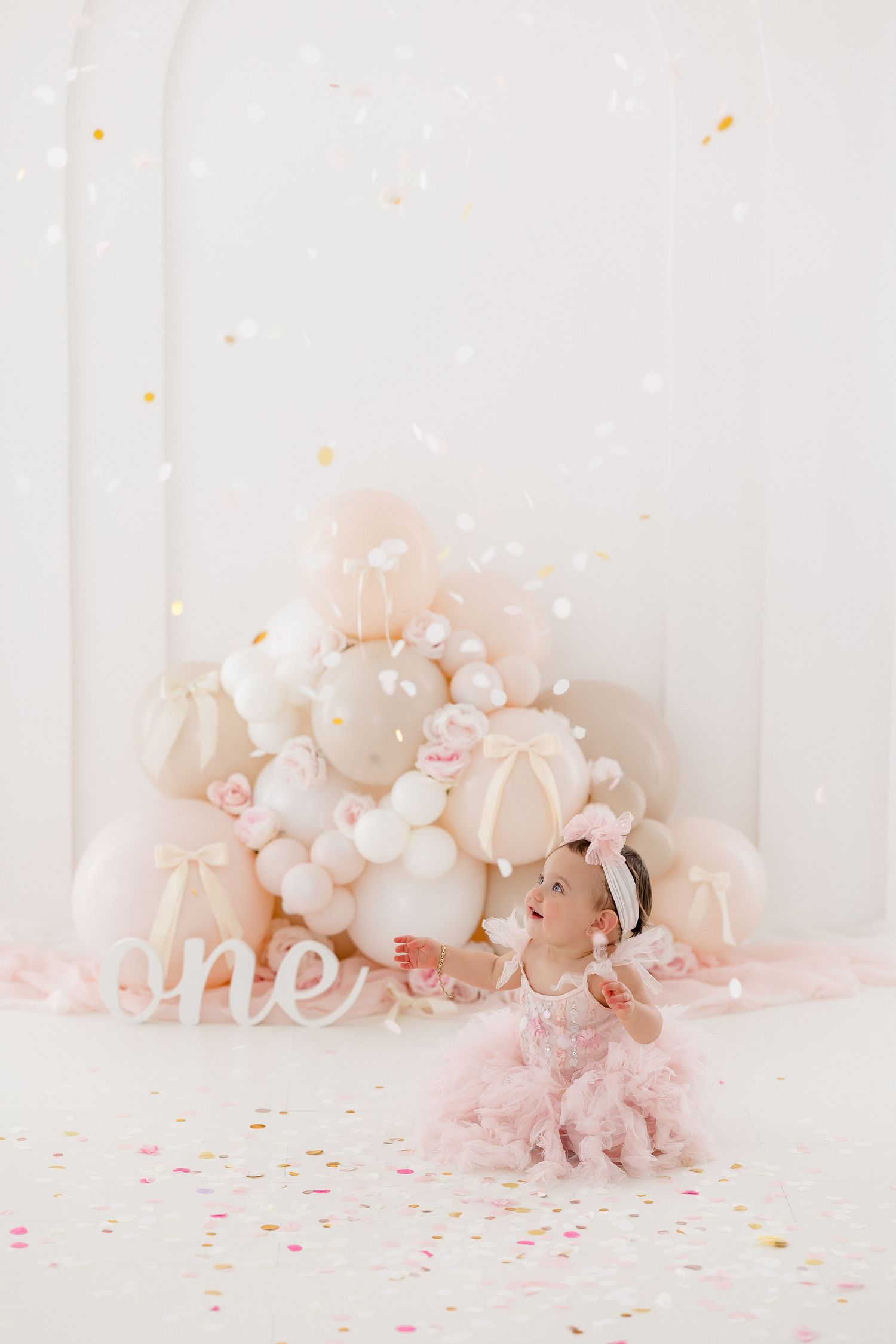 Pink and white balloon arch decorates a delicate nursery setting with scattered confetti on a light background.