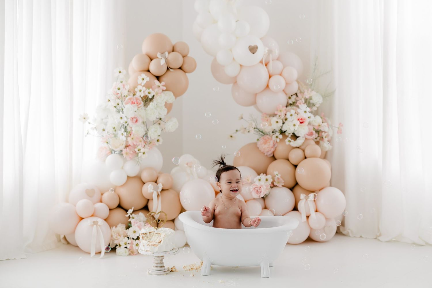 Pink and peach balloon arch with floral arrangements and white curtains creates an elegant party backdrop setting.