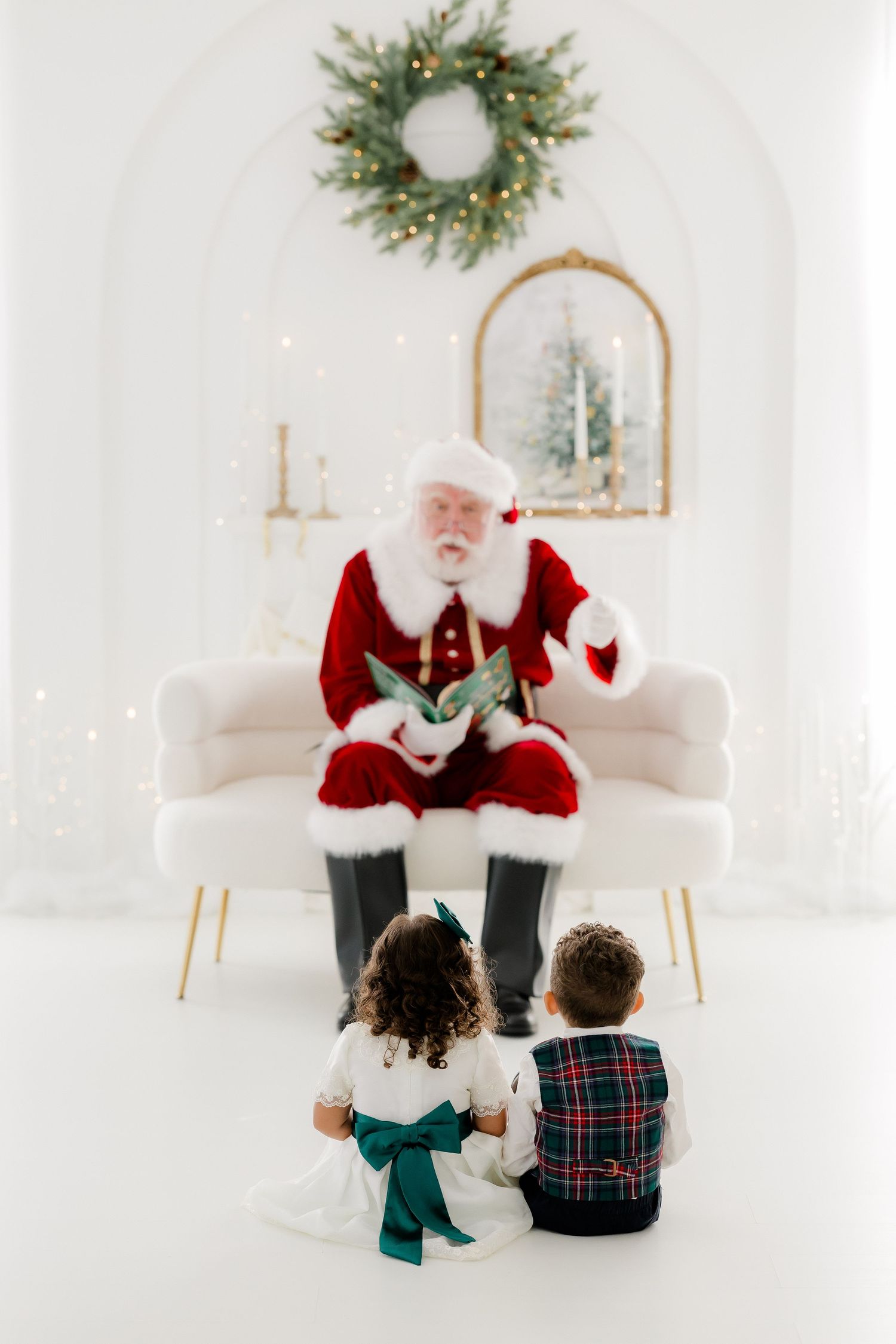 Santa Claus reads a book to two young children sitting on the floor in a bright white Christmas-decorated room.