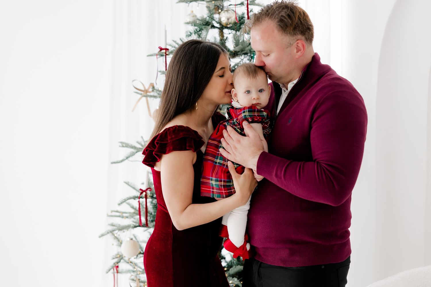 Family wearing burgundy outfits shares intimate moment by Christmas tree.