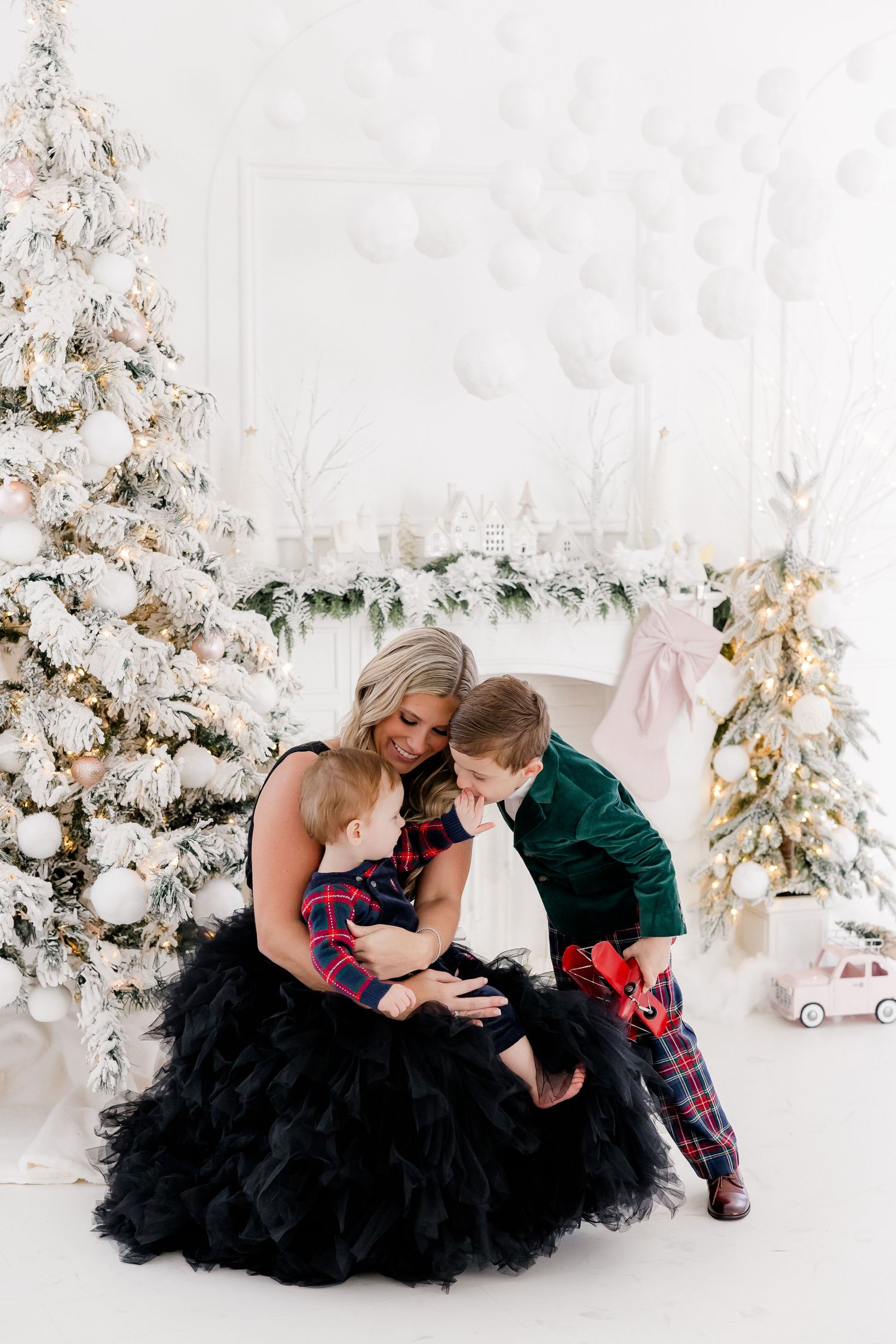Holiday photoshoot featuring black tulle dress and plaid accents against white Christmas backdrop.