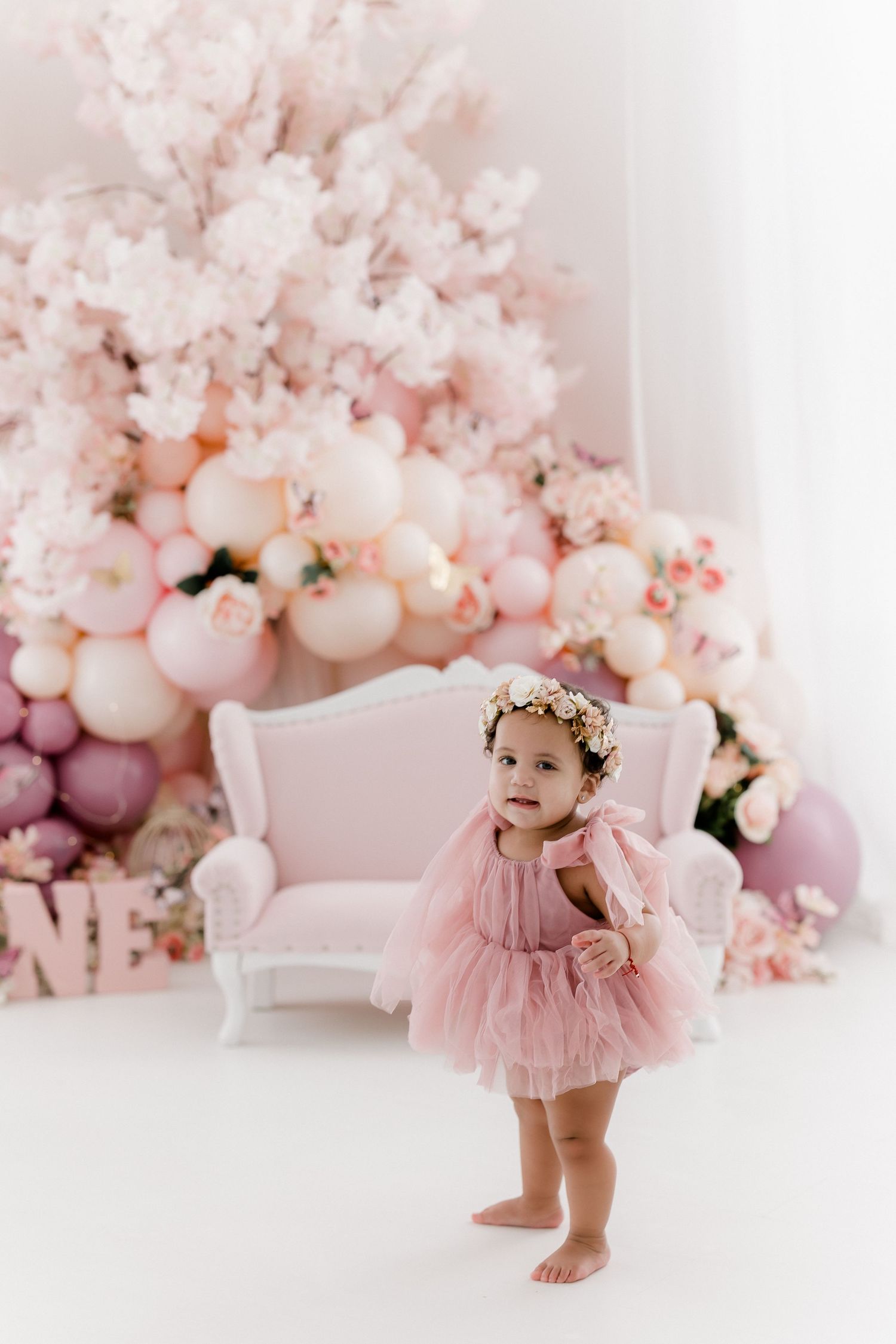 Baby in pink tulle dress playing against backdrop of pink balloons and flowers.