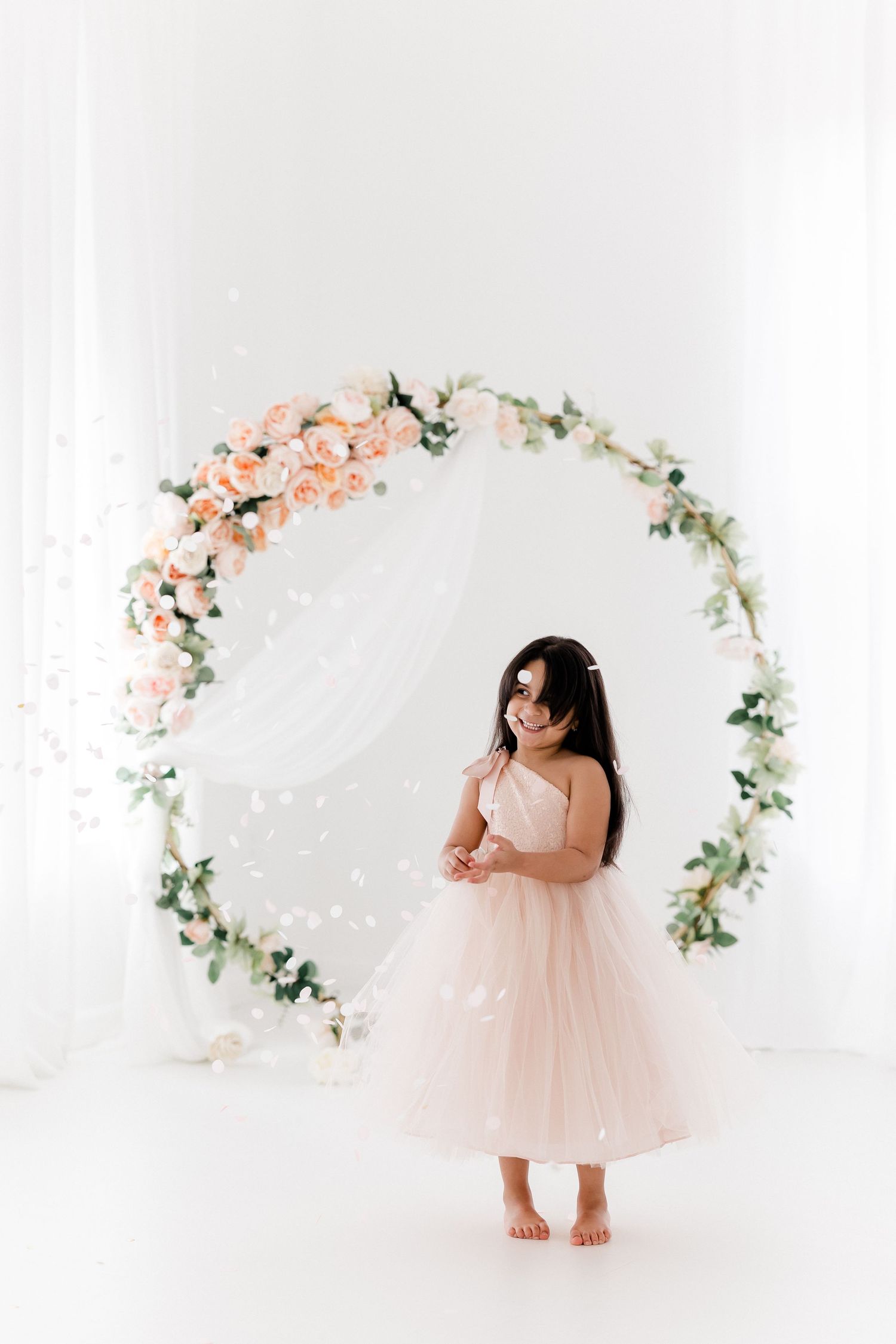 A child in a pink tulle dress twirls and plays in a dreamy white studio setting with floral arch decorations.