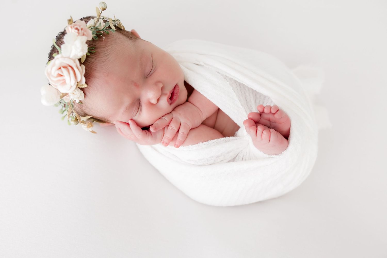 A sleeping newborn wrapped in white fabric wearing a delicate floral crown on a light background.