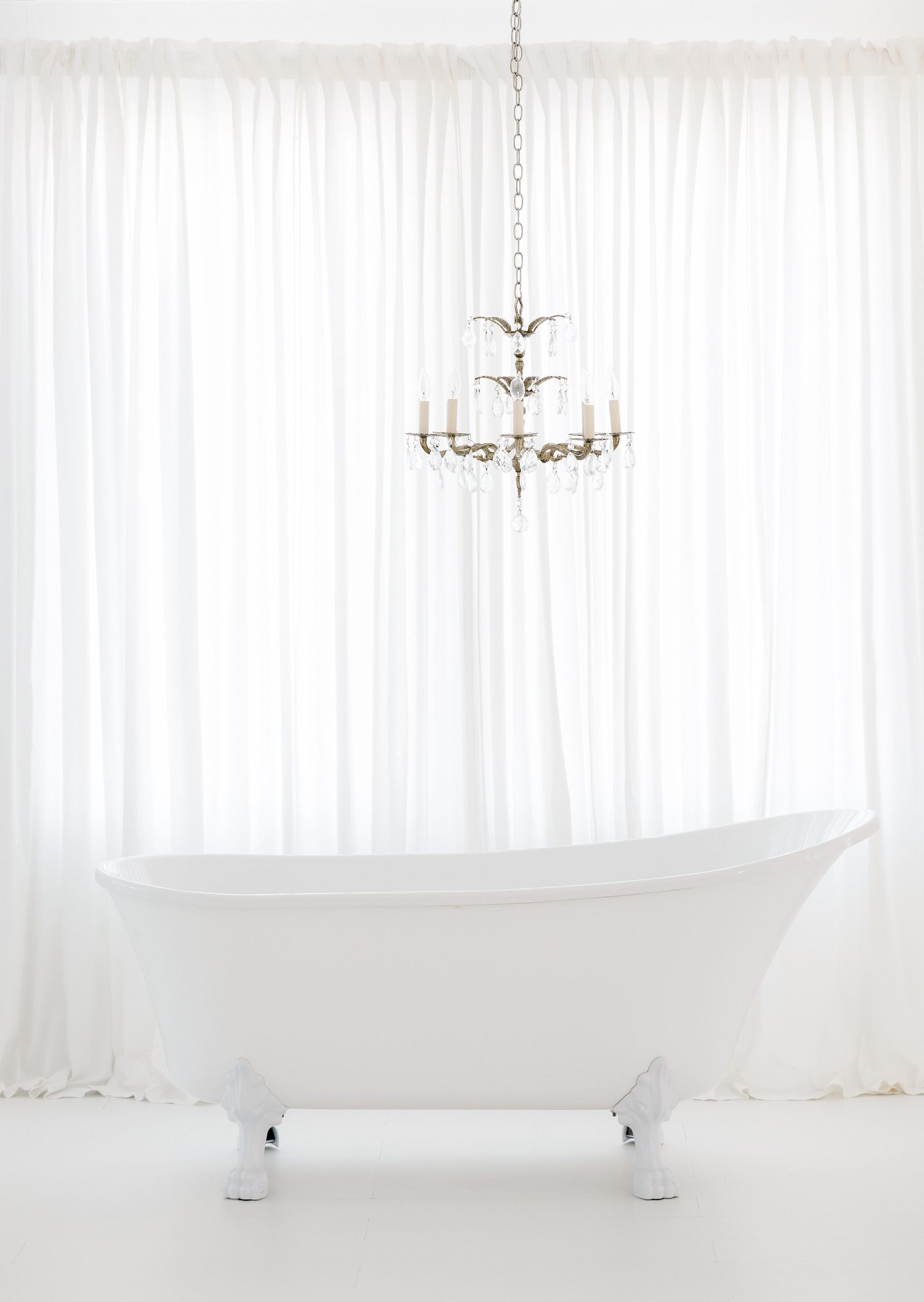 Elegant white bathtub in minimalist bathroom with sheer curtains and crystal chandelier creating a luxurious atmosphere.