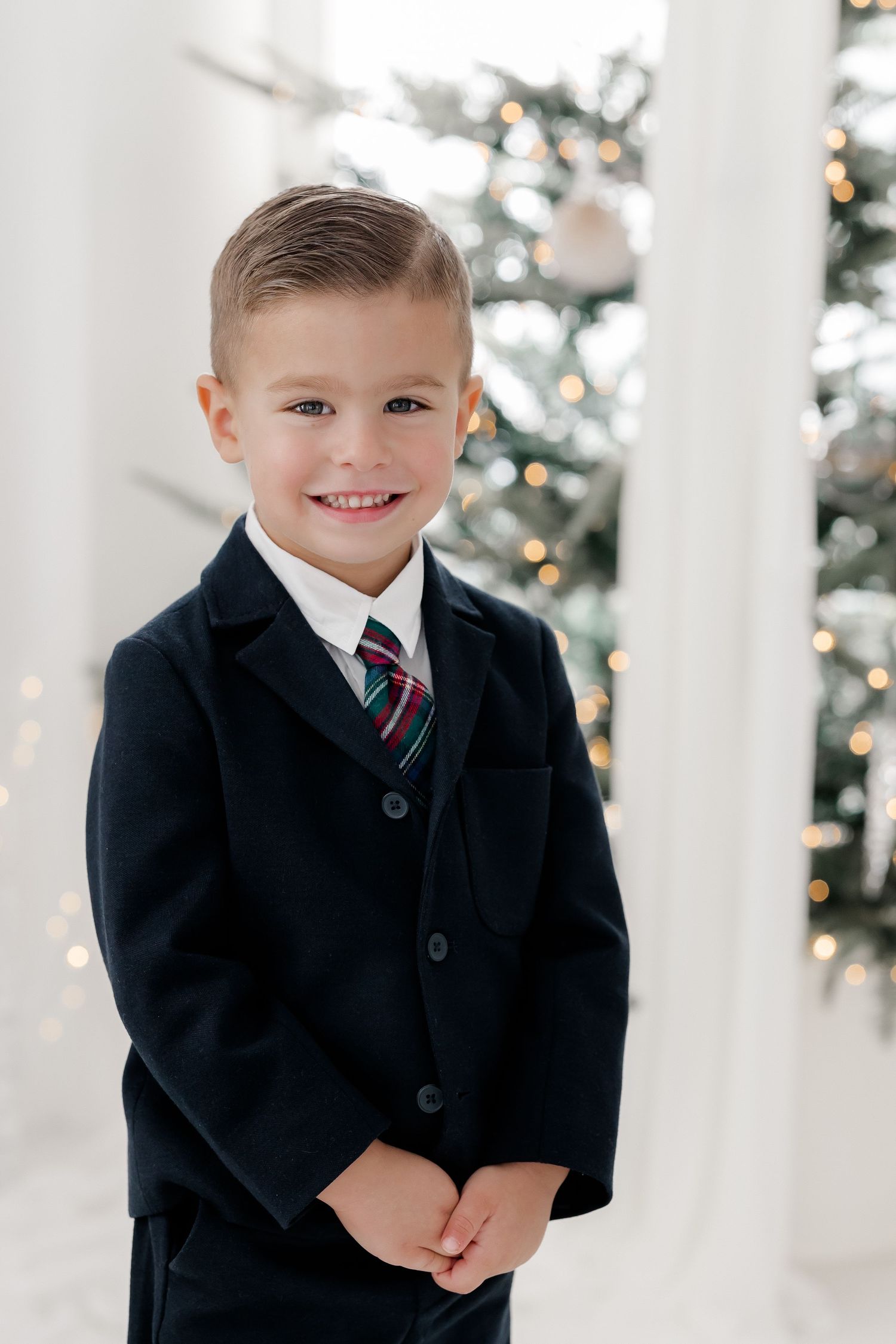 Young boy in navy suit and striped tie smiling in front of decorated Christmas tree with white columns.