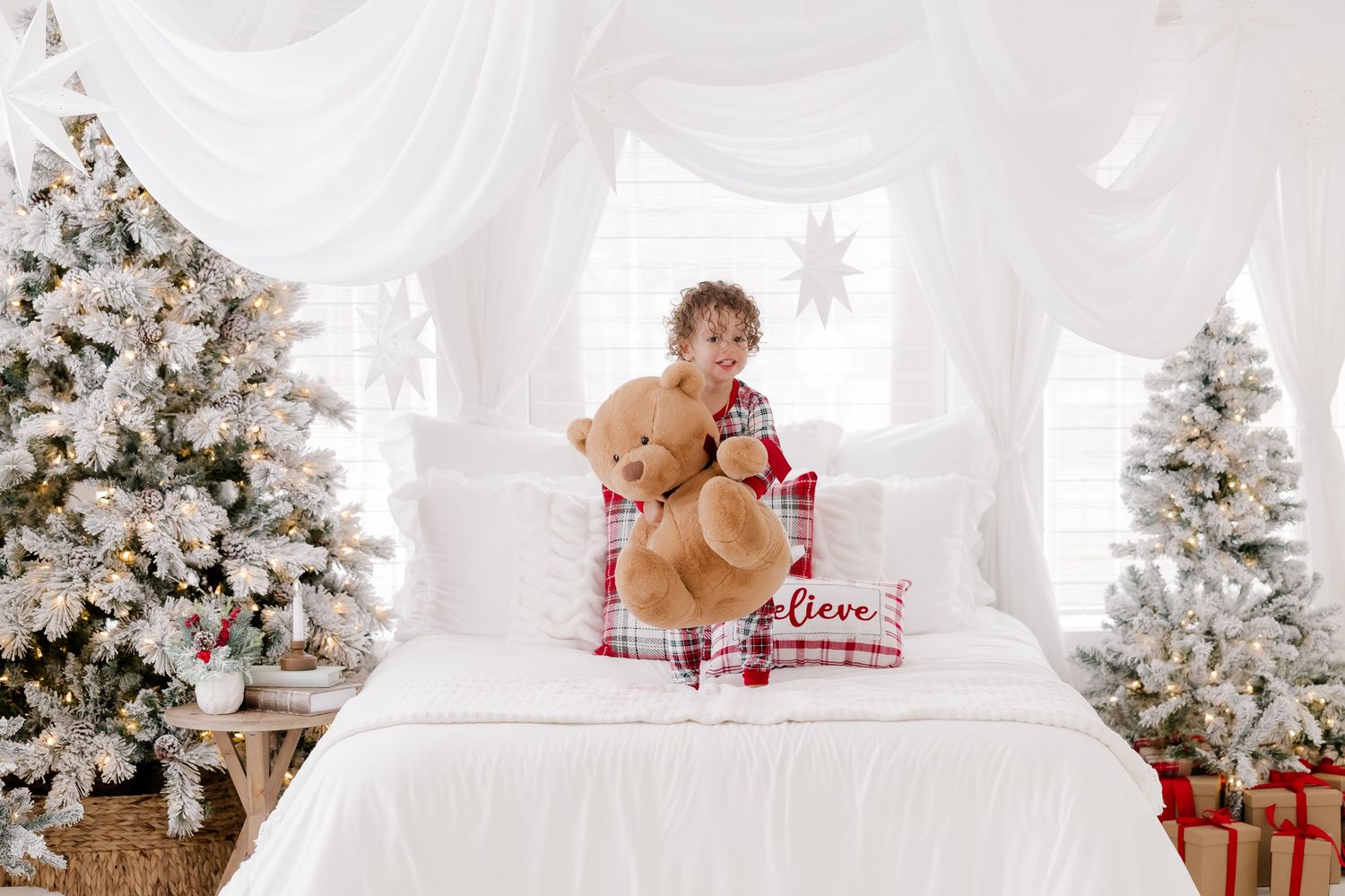 Child in Christmas pajamas sitting on white bed holding teddy bear, surrounded by flocked Christmas trees and gifts.
