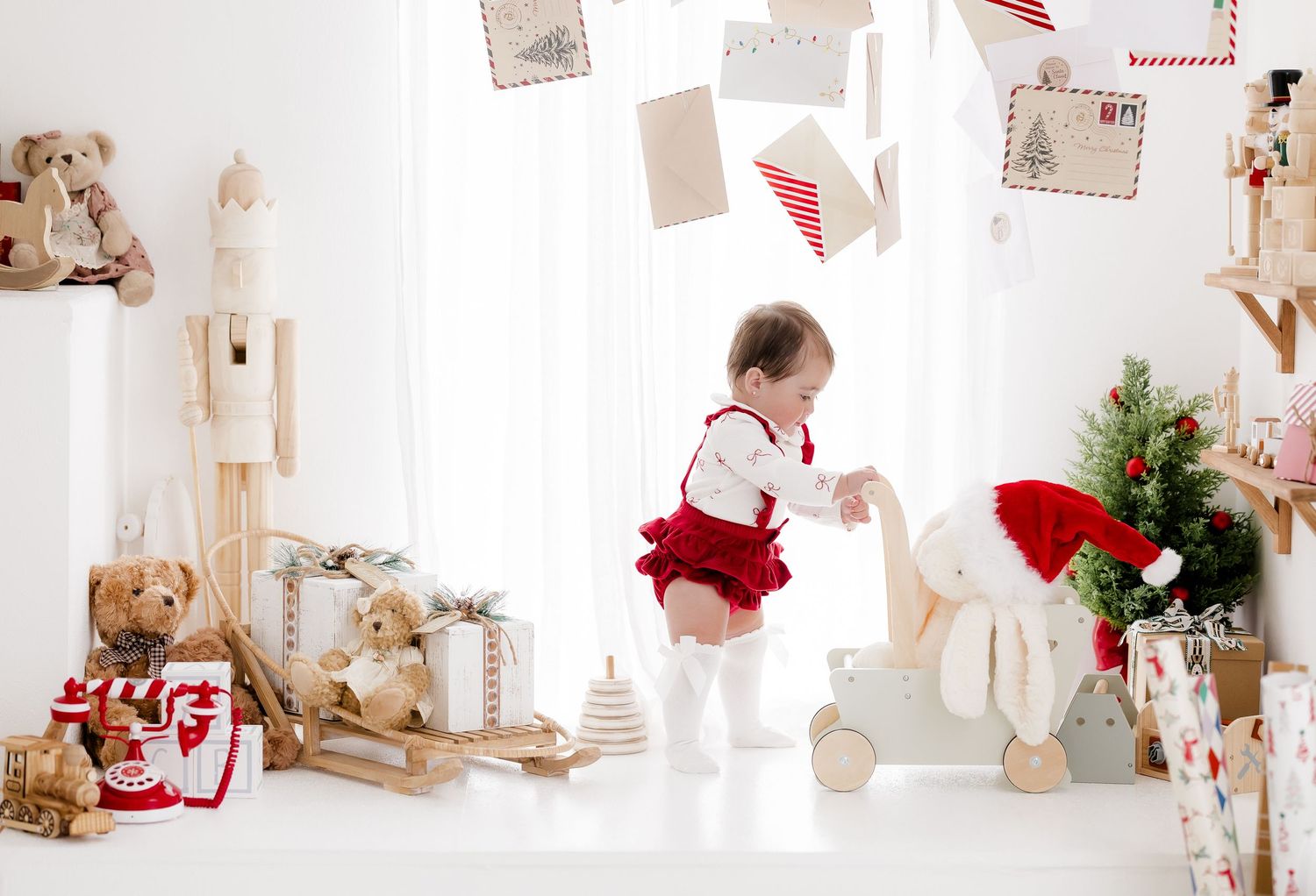 Toddler in red Christmas outfit playing with toys in a festive white room with teddy bears and holiday decorations.
