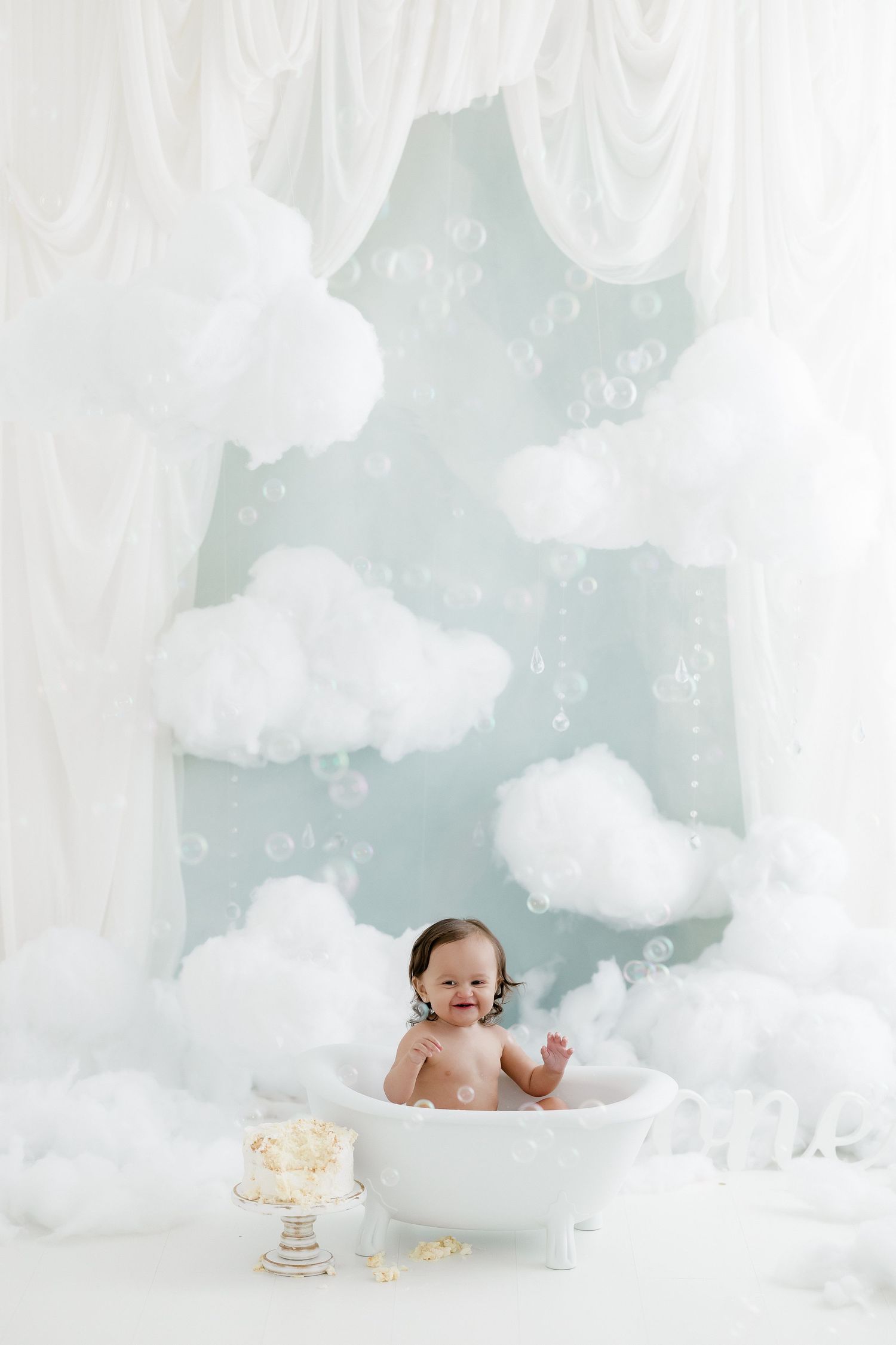 Baby sitting in a small white bathtub surrounded by fluffy clouds, bubbles, and white curtains in a dreamy studio setup.