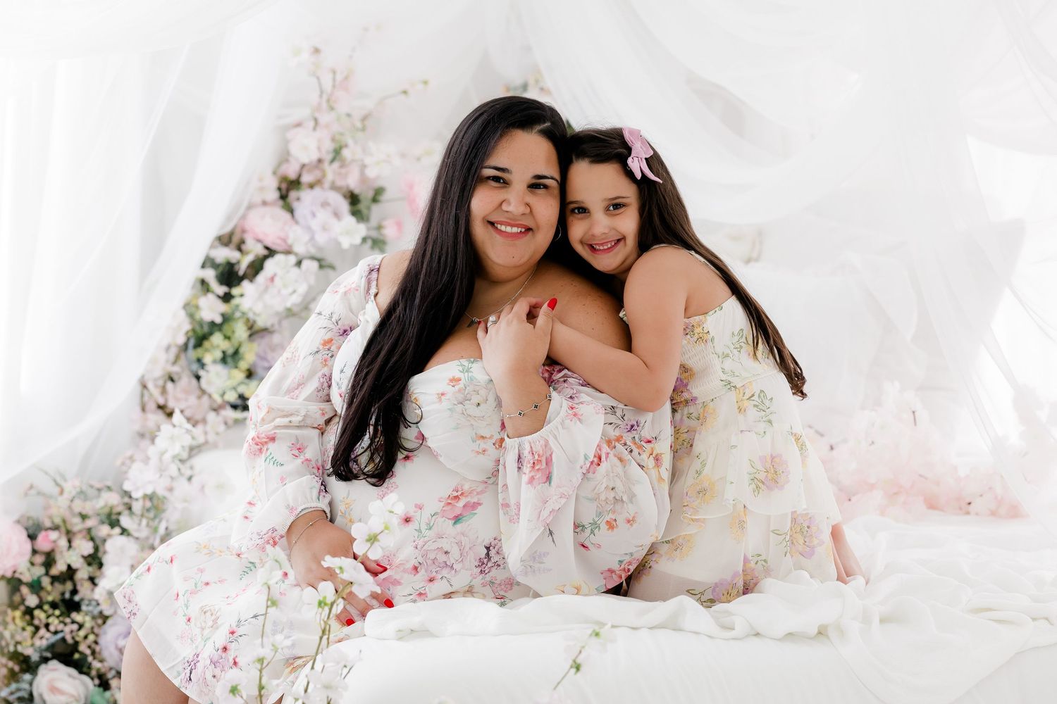 Two people in white floral outfits pose together in a romantic studio setting with soft pink floral decorations.