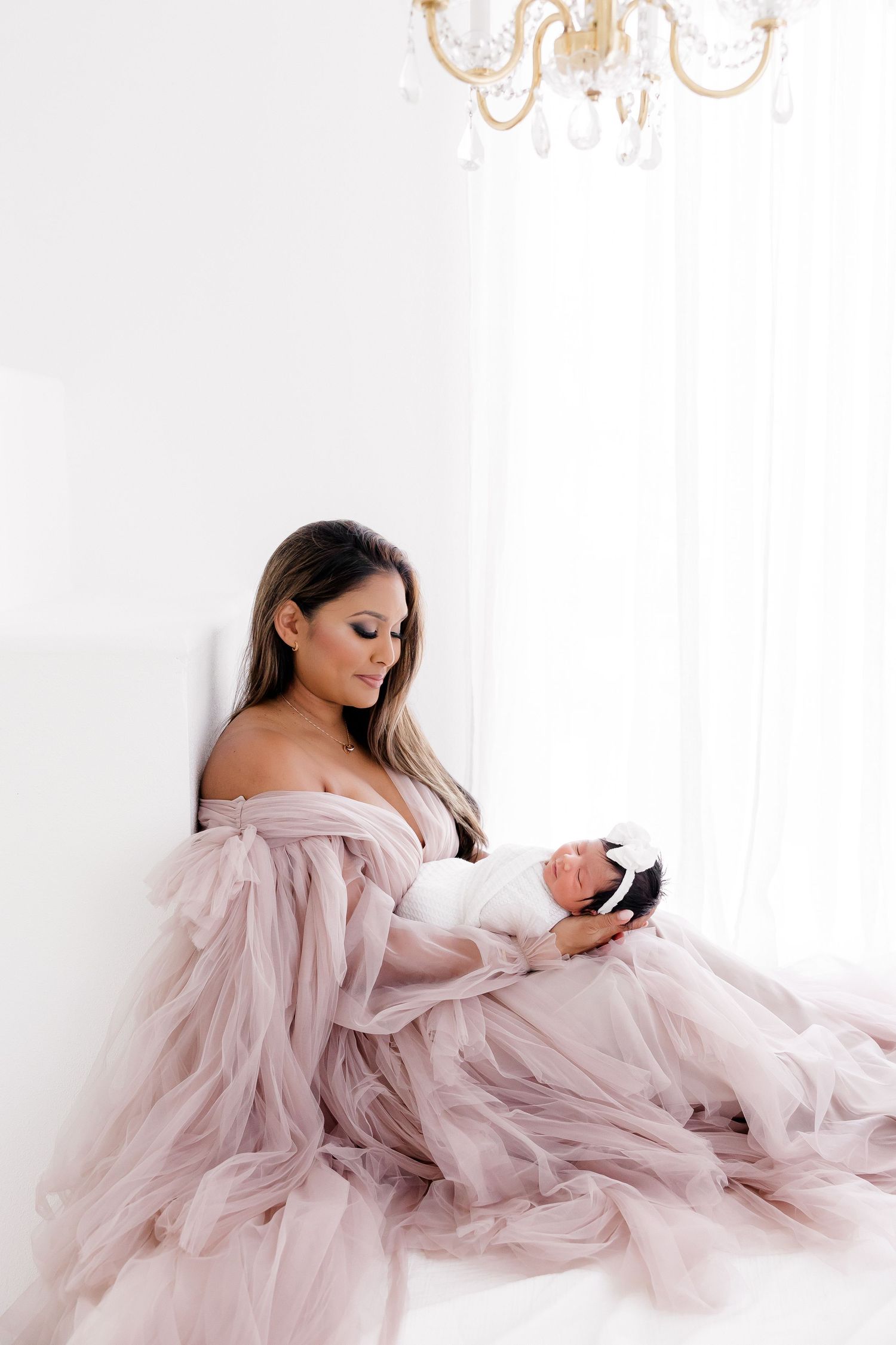 Mother in flowing pink dress holds newborn beneath elegant chandelier.