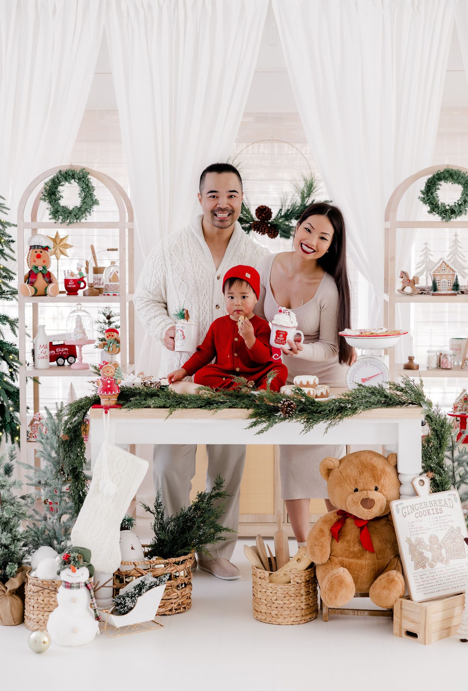 Holiday family photoshoot scene with decorative Christmas trees, wreaths, and a teddy bear in a bright white studio setting.
