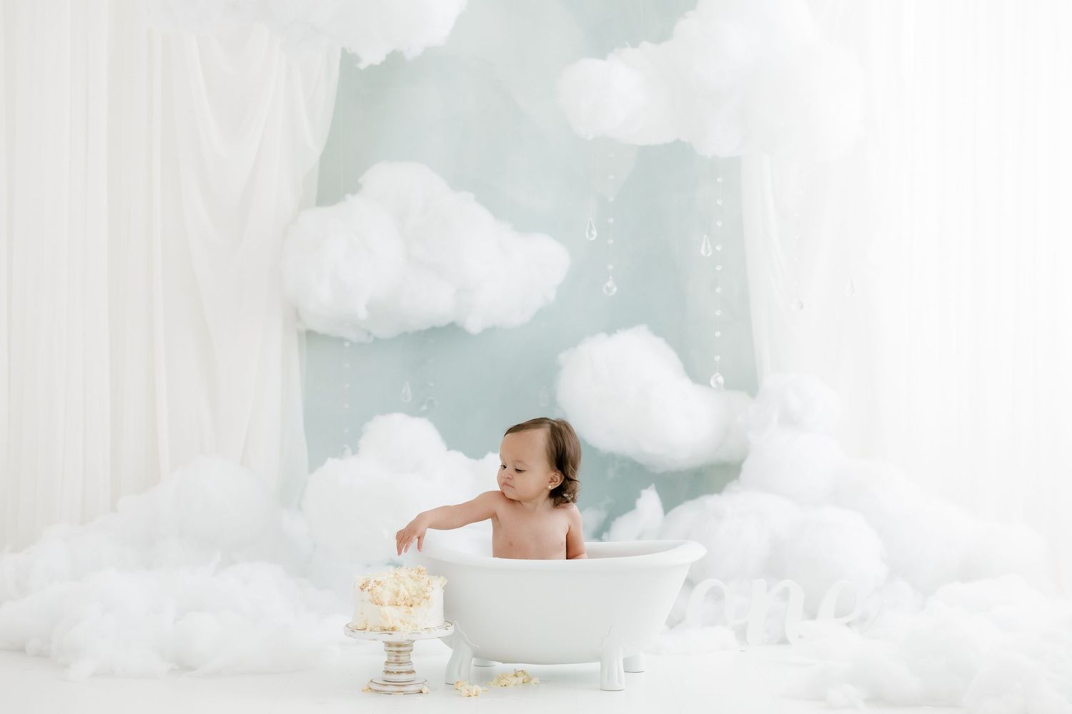 Baby sitting in a white bathtub surrounded by fluffy clouds and white balloons in a dreamy studio setting.