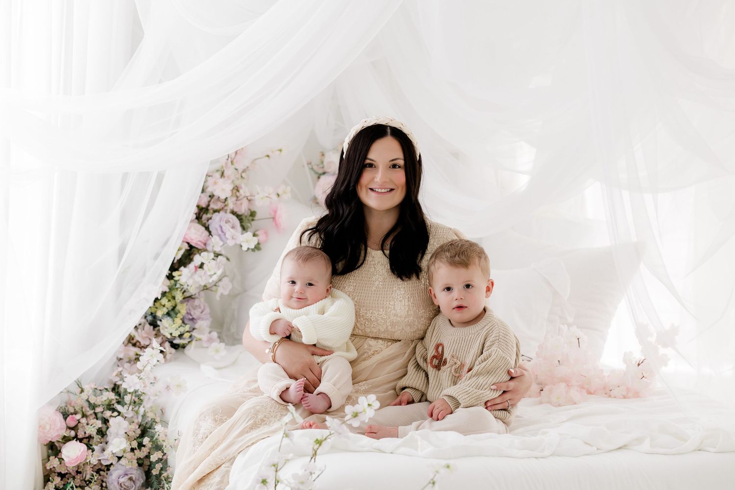 A mother and baby pose together on a white bed surrounded by delicate pink floral arrangements.