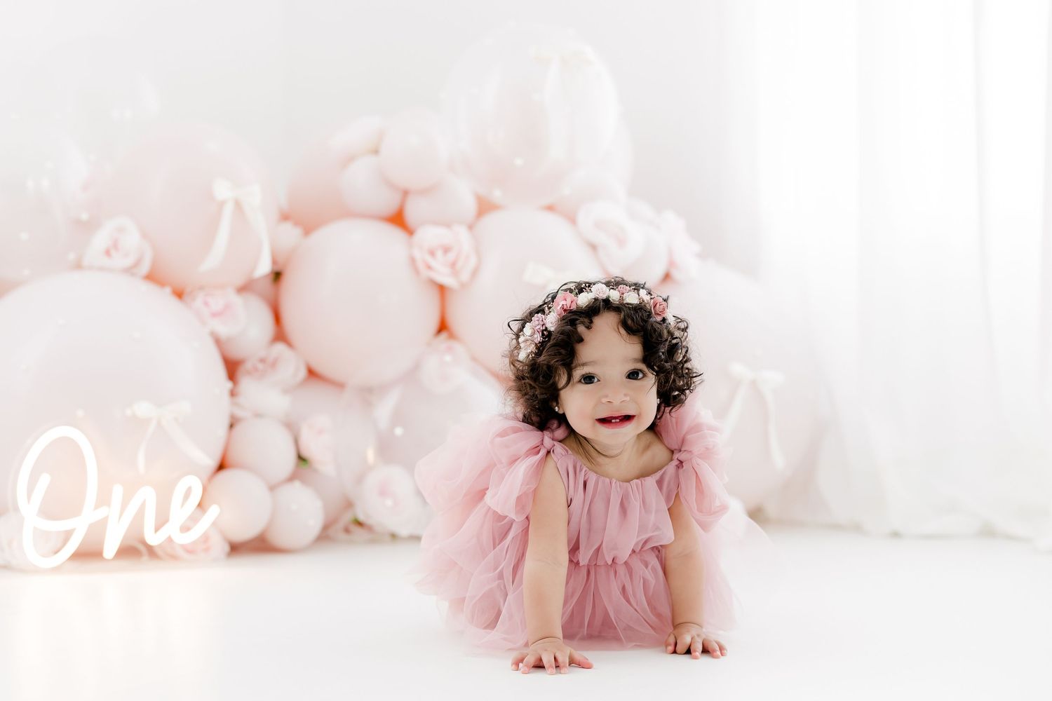Child in pink dress lies on white floor surrounded by soft pink balloons.