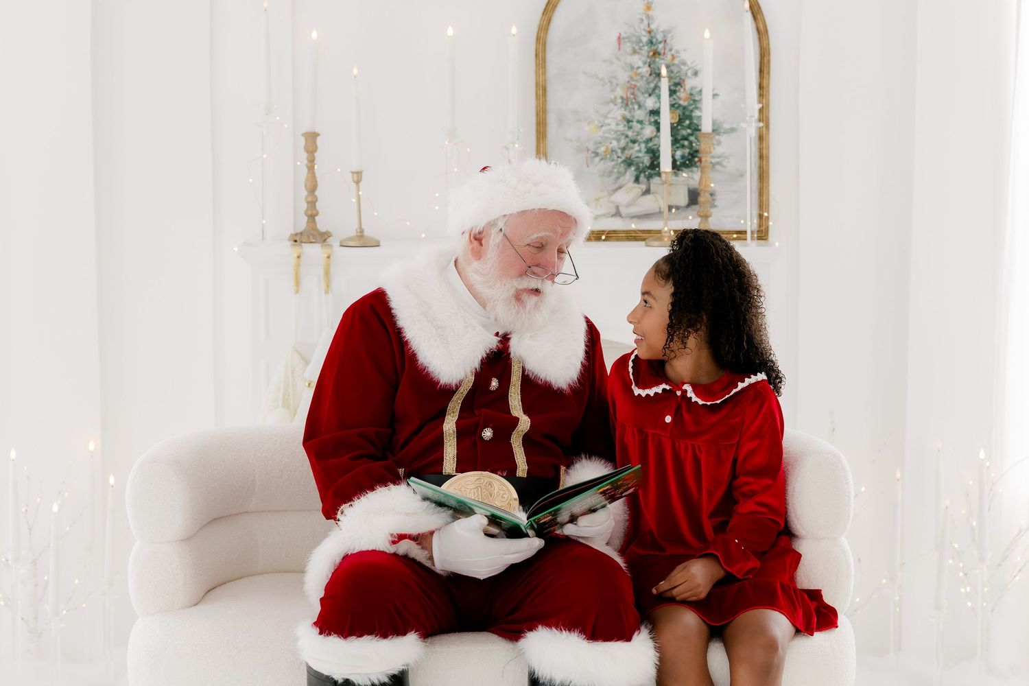 Santa Claus reading a book with a young girl in a red dress on a white couch in a festive setting.