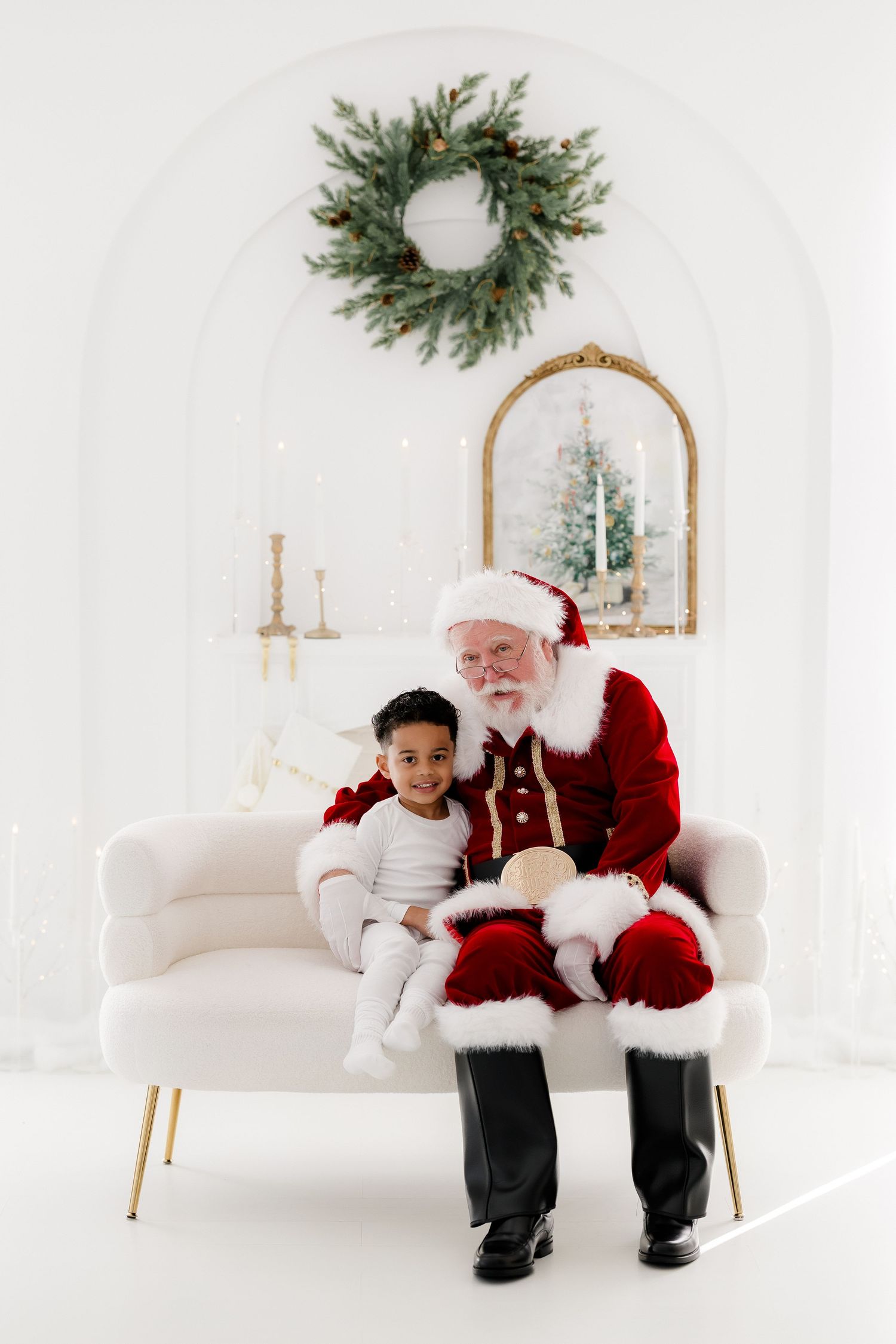 A child sits on Santa Claus's lap on a white velvet sofa in a bright holiday studio with a wreath and mirror.