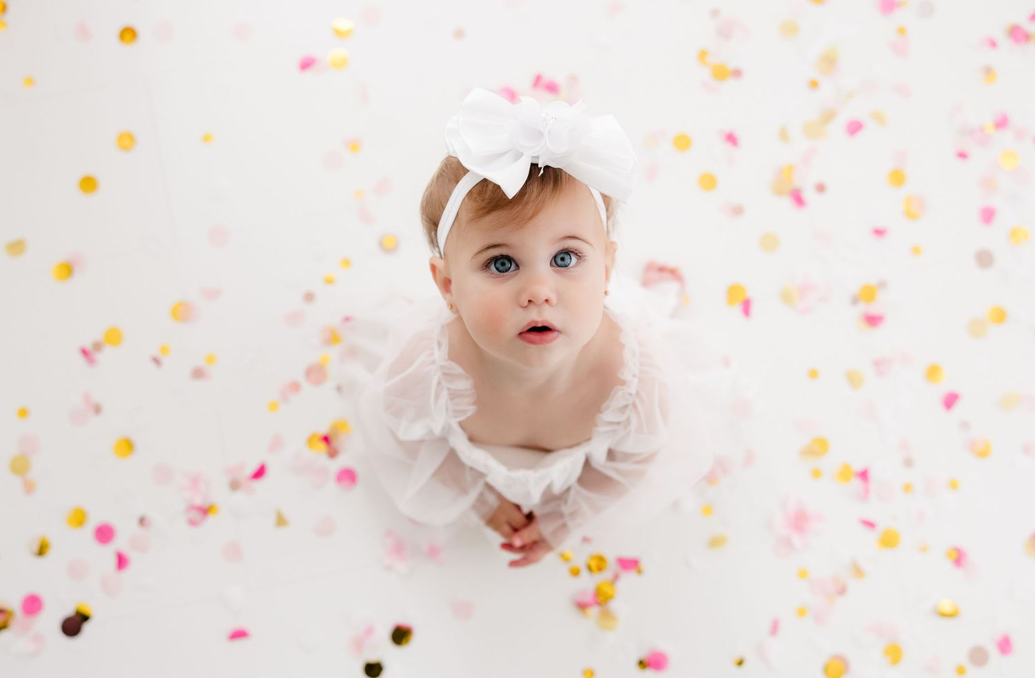 Baby in white dress surrounded by colorful confetti on white background.
