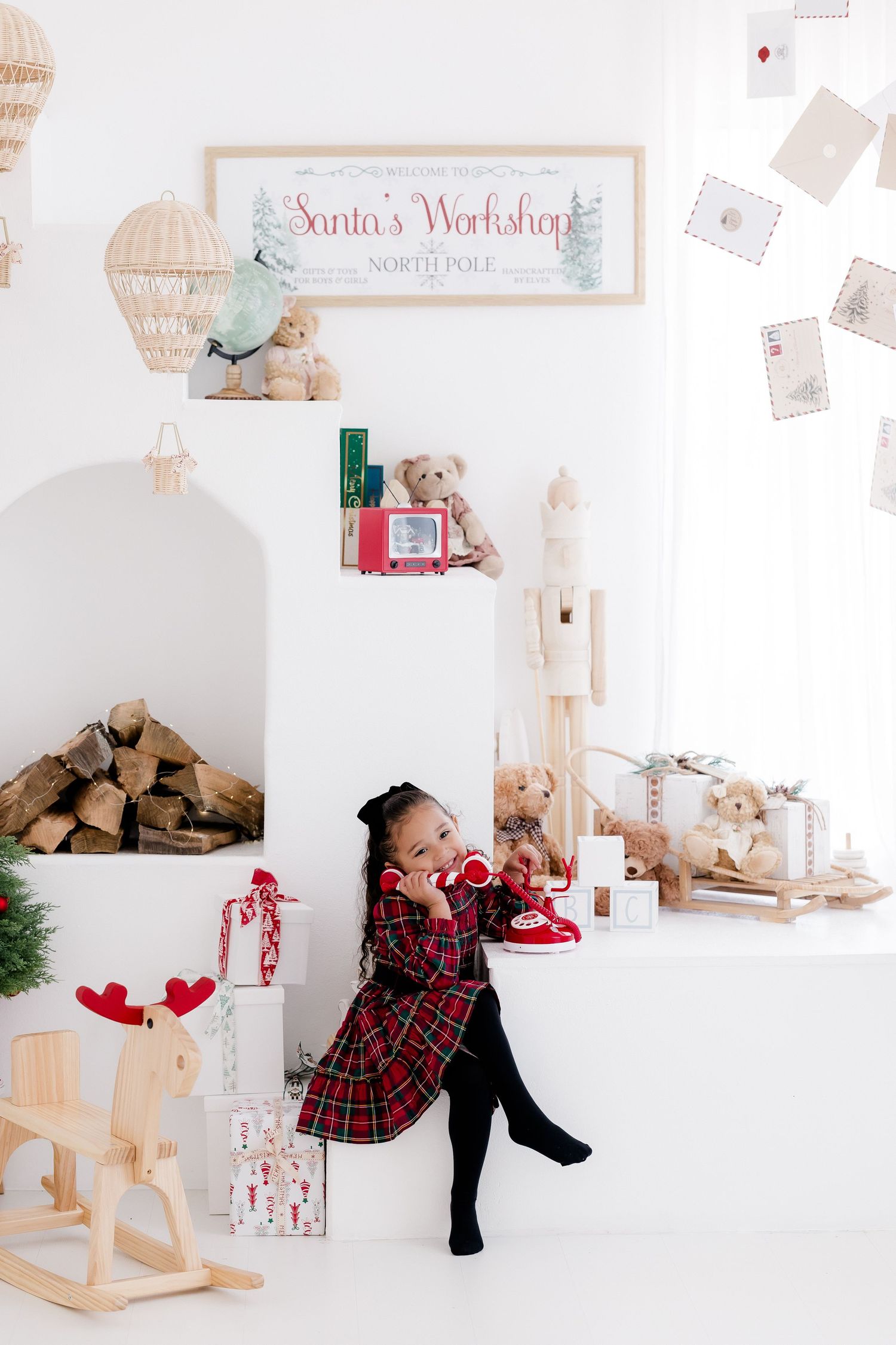 Young girl in plaid dress poses by Christmas decorations near Santa's Workshop sign with toys and gifts.