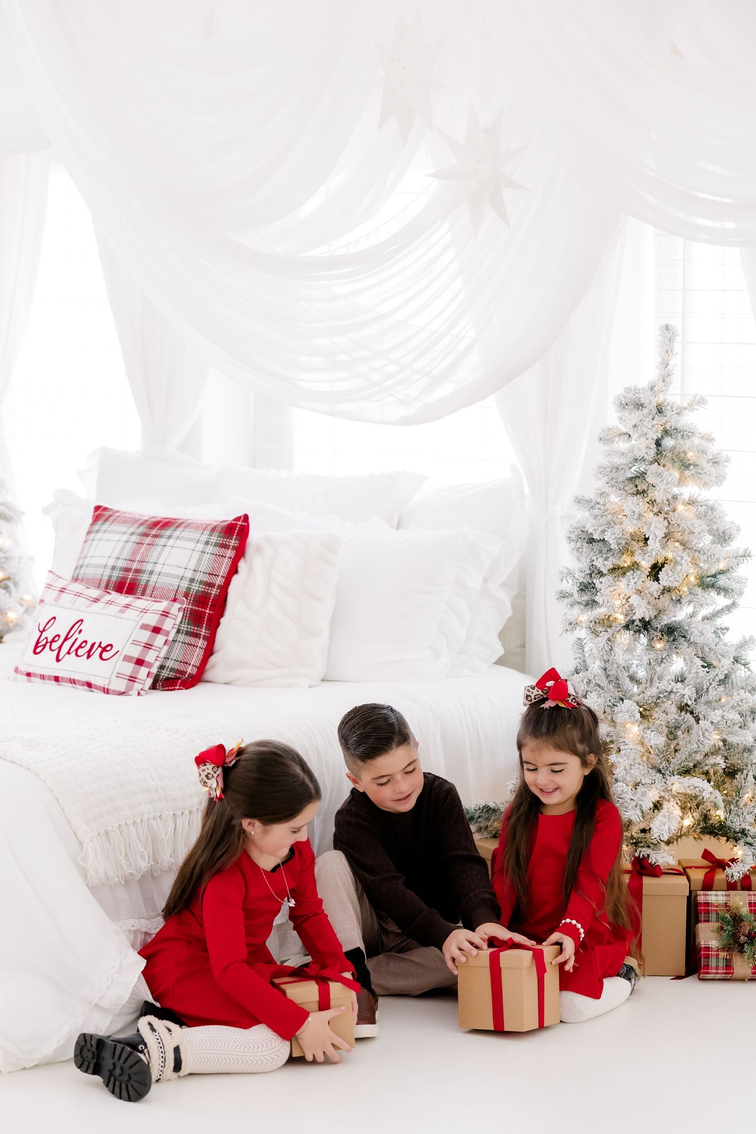 Three children in red outfits open Christmas gifts beside a decorated tree in a white holiday bedroom.