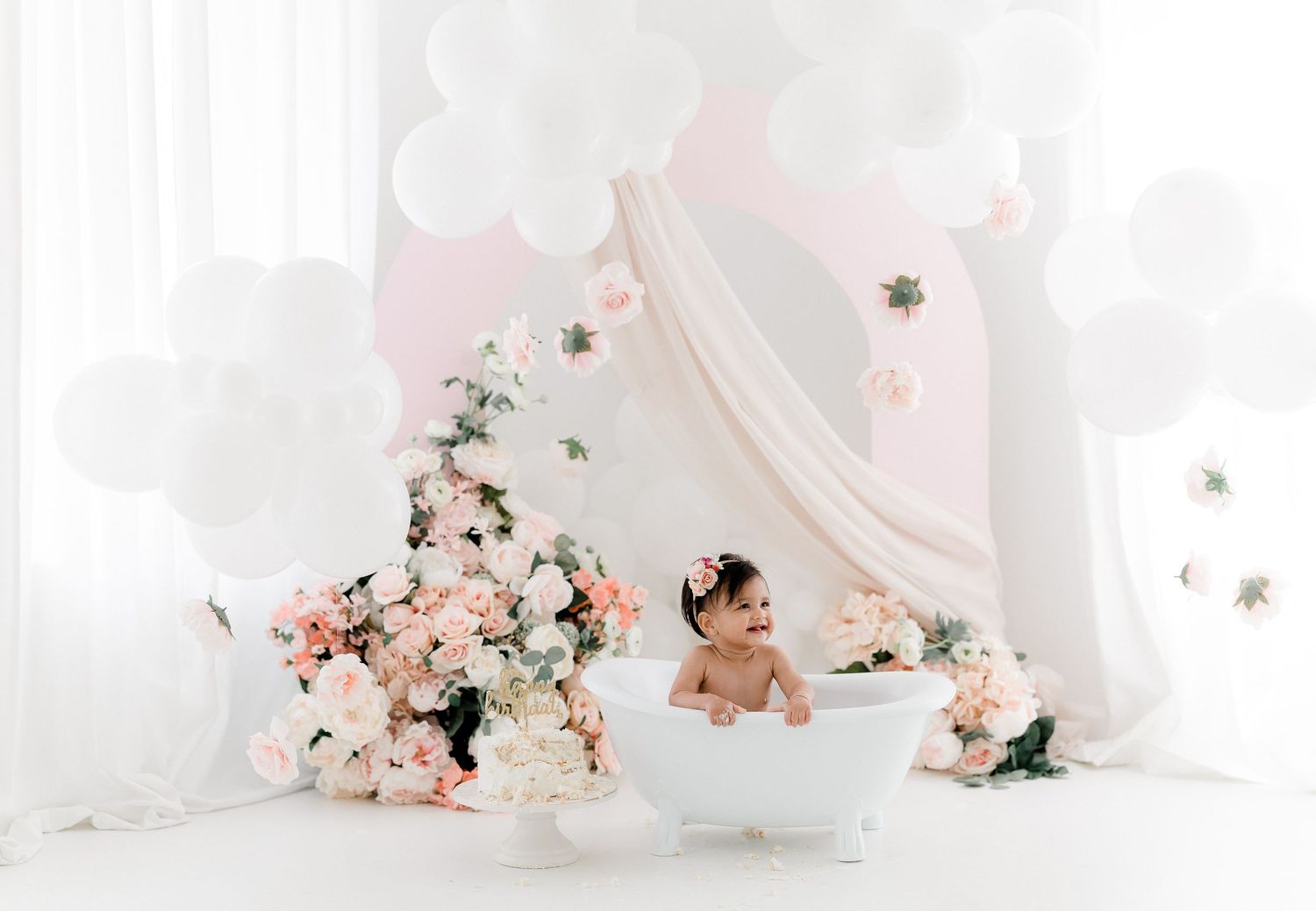 A white bathtub decorated with pink and white flowers sits against a dreamy white backdrop with flowing curtains.