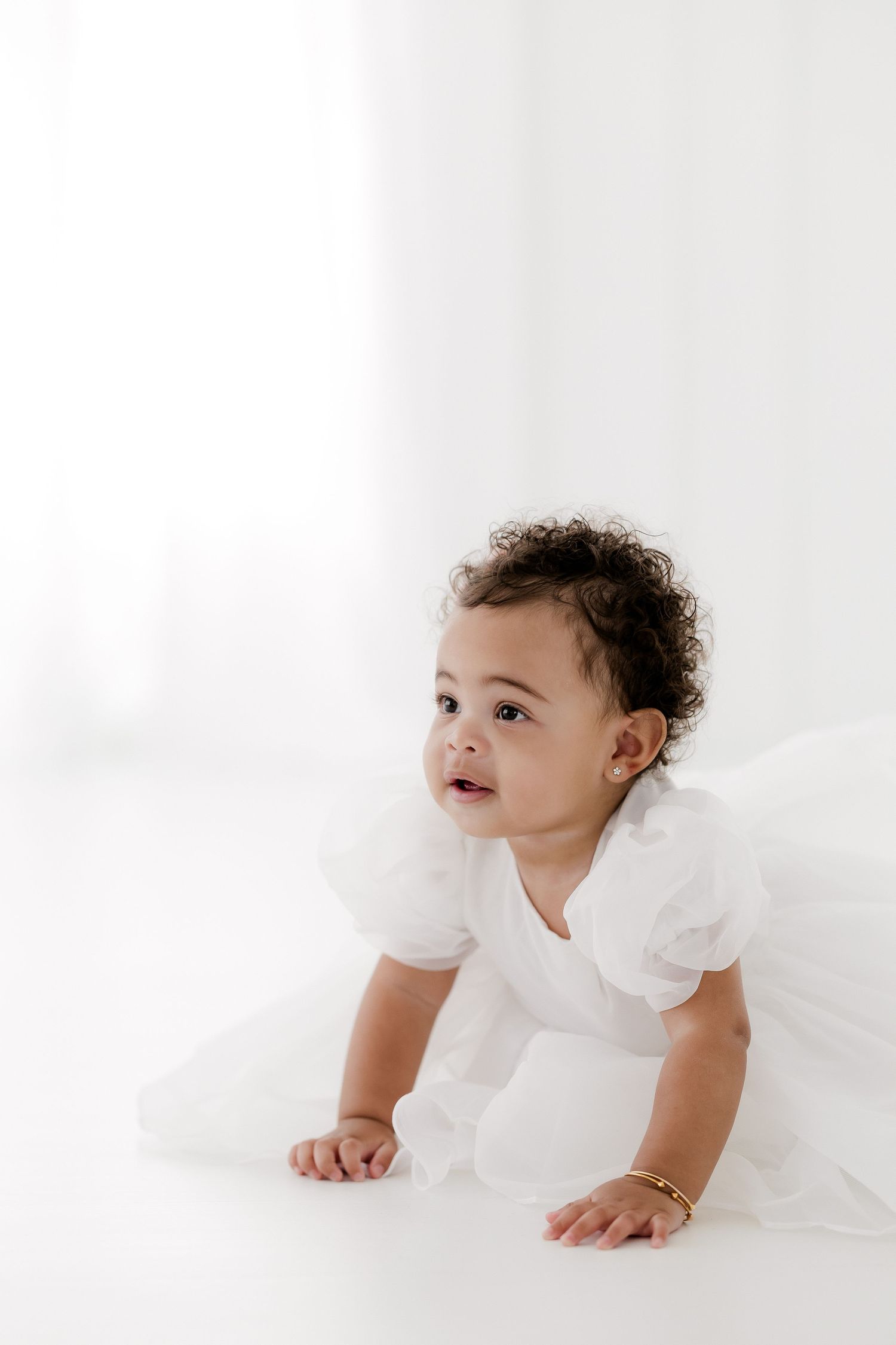 Baby in white dress with curly hair crawls on white backdrop during minimalist portrait session.