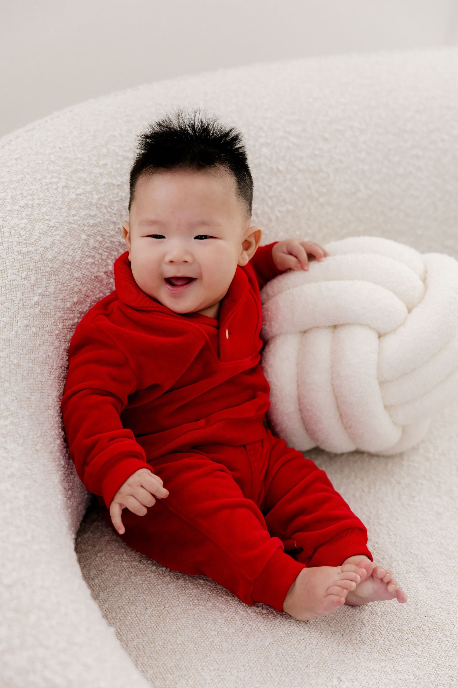 Baby in red outfit lying on white textured surface with festive holiday backdrop.