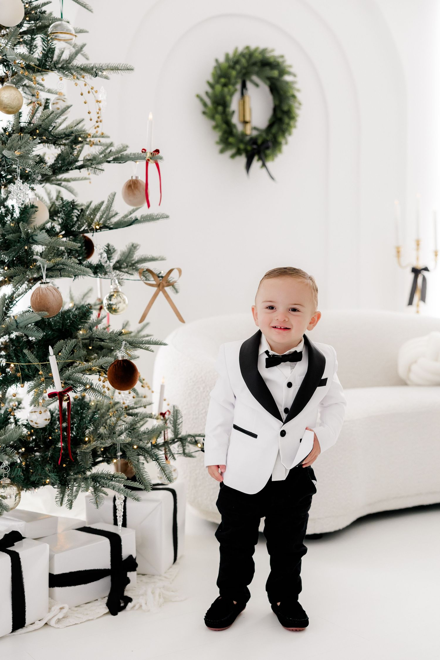 Young formal attire near decorated Christmas tree in elegant white room.