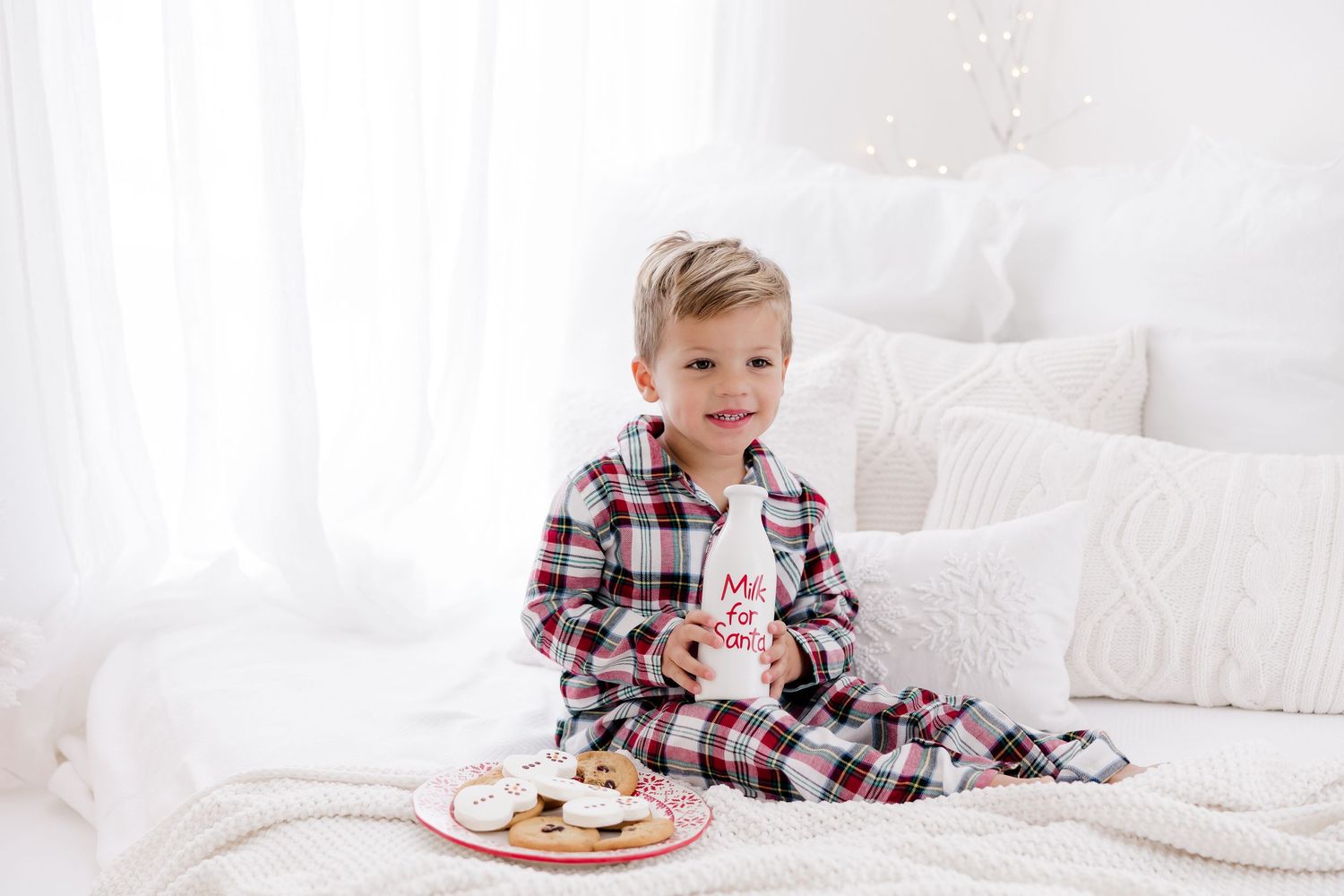 Child in plaid pajamas enjoys milk and cookies on white bedding during holiday season.